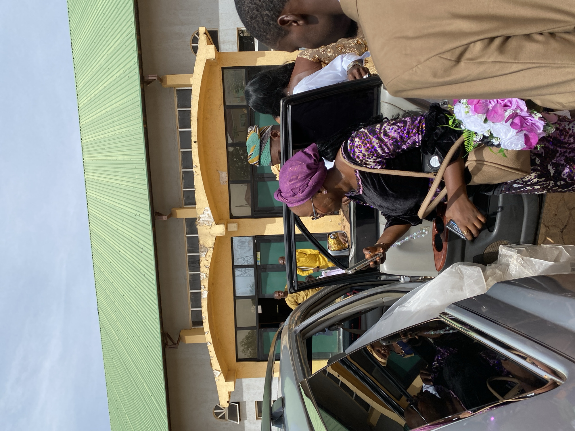 In Kuru Karama, Nigeria, a woman wearing a purple headwrap and sequined top exits a silver car with a bouquet of flowers. She is holding a phone in her right hand. The building behind has a green roof and yellow exterior with arched windows. Other people stand around the car, including a man in a tan suit and a woman in a white dress. A man in a yellow shirt can be seen in the building's doorway.