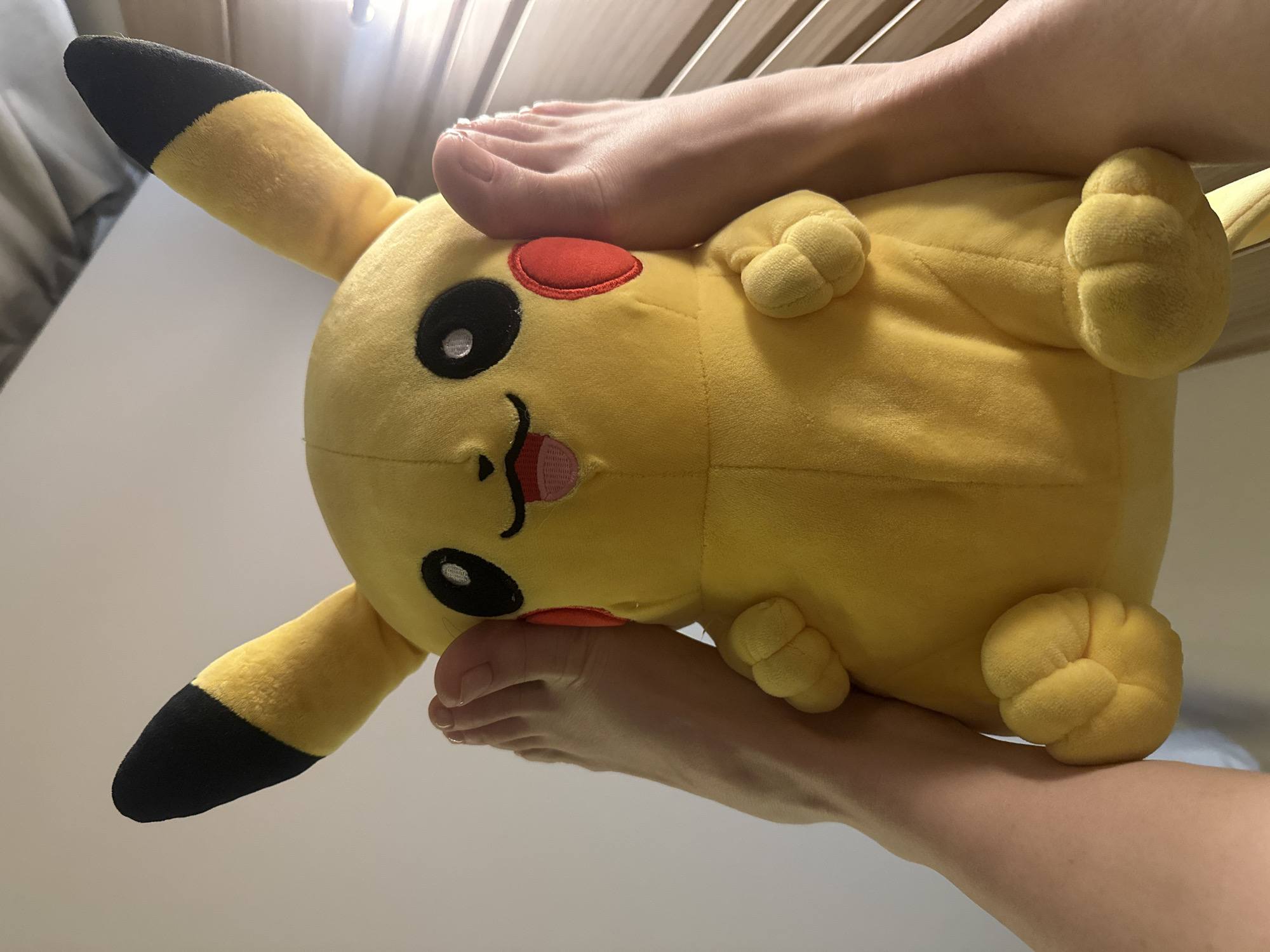 A plush yellow Pikachu toy is partially obscured by a person's bare feet. The person's feet are positioned on either side of the Pikachu. The toy is lying on a flat surface, possibly a bed or table. The background is blurred and shows elements of a room. There is no evidence to place this image in Votkinsk, Russia.