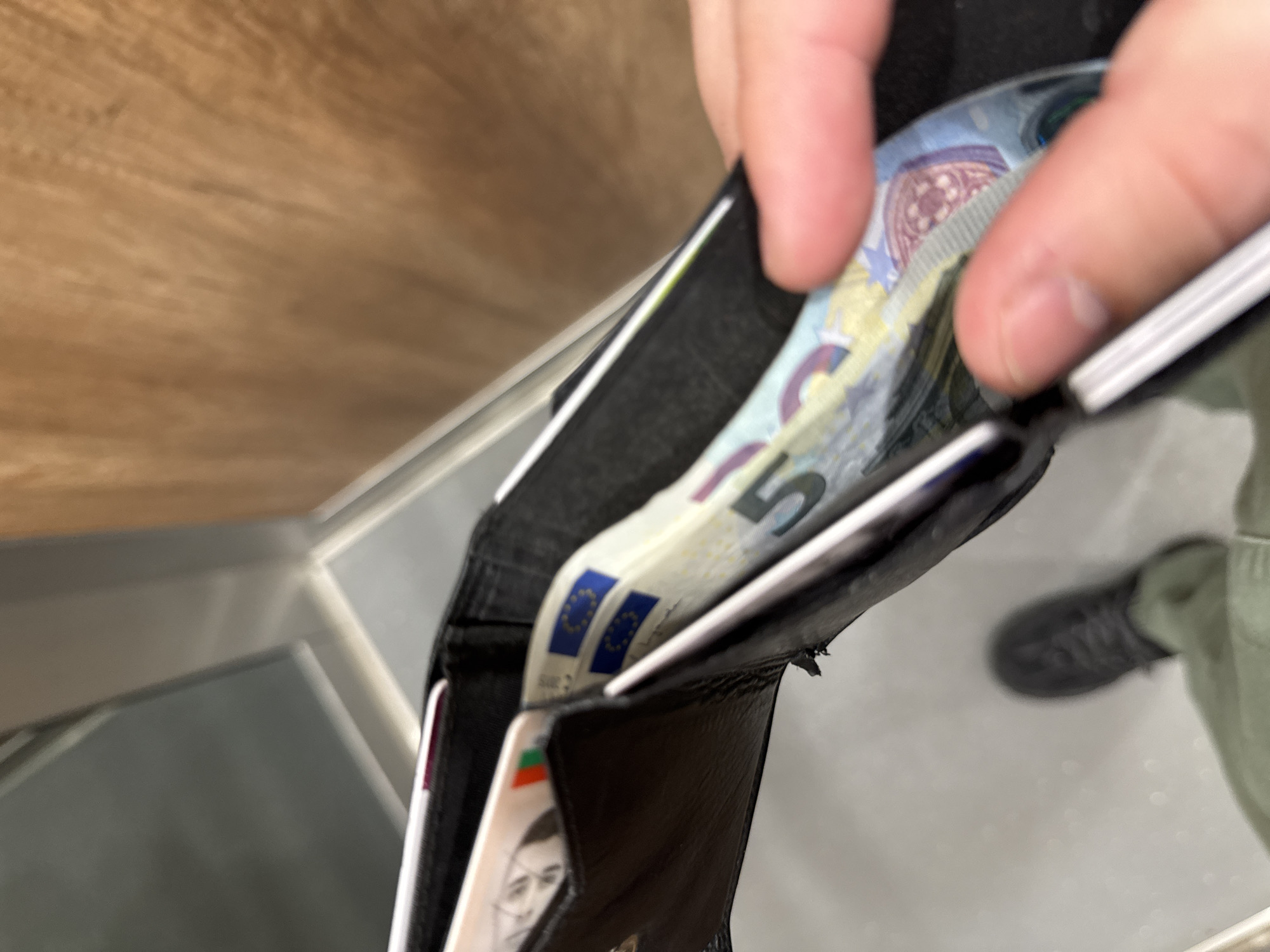 The image depicts a close-up, high-angle shot into an open black leather wallet. The wallet is held open with the thumb and forefinger of a person's right hand, revealing several euro banknotes inside. The visible denominations include a 5 euro note. There is a card with a picture of a man on it, and a European Union flag is visible on the banknotes. The lower right corner of the image shows the person's leg, including a dark-colored shoe and light green pants. The background consists of a mix of surfaces, including a light-colored wood-grain surface and a silver metal railing. The photograph was taken indoors, and the location is uncertain. The information about being in Sofia, Bulgaria is not directly verifiable from the image itself.