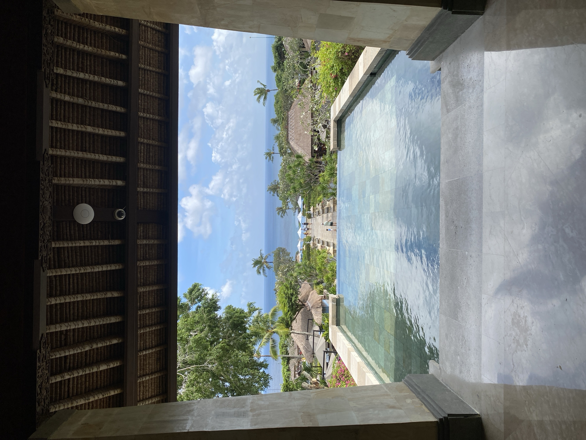 The image depicts a view from an indoor structure, likely a villa or hotel, towards an outdoor scene in Kuta Selatan, Indonesia. The composition is framed by a thatched roof ceiling and a stone structure encompassing a swimming pool that seamlessly blends with the ocean view.
The central view reveals a coastal landscape under a blue sky with scattered clouds. Vegetation, including trees and palm trees, covers the area, with traditional Balinese-style structures (possibly bungalows or villas) interspersed among the foliage. In the distance, the ocean stretches towards the horizon.
To the right of the frame, a portion of a swimming pool is visible. The water surface reflects the sky and the surrounding environment. The foreground structure of the villa or hotel has stone steps leading to the pool area.
No individuals are clearly visible in the frame. The image captures a serene scene, emphasizing the architecture and natural environment of the location.