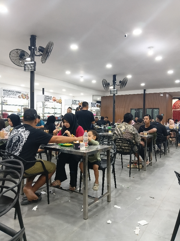 The image depicts an indoor, brightly lit food court or casual restaurant in Seumadam, Indonesia. Numerous patrons are seated at rectangular, metal-framed tables with black chairs, engaged in dining and socializing.
The environment features a white ceiling with multiple recessed LED lights and several black poles supporting dual-head industrial ceiling fans. The floor is light grey concrete or large tiles, with scattered white paper napkins. Signs in Indonesian advertising "WARUNG BU MINAH" and "DIMSUM PEMPEK DESE" are visible on the white walls behind glass display cases in the left background. A dark brown wooden panel wall occupies the right side of the dining area. A black security camera is mounted on the ceiling in the mid-ground.
In the foreground, a man in a black t-shirt with a white graphic print on the back and olive green shorts, wearing flip-flops, is seated with his back to the camera, reaching towards a plate. Opposite him, a woman in a red shirt and black hijab is actively eating from a plate with a fork. To her right, a young child in a light green t-shirt and dark shorts sits, looking towards the right, with a plastic water bottle on the table. Other individuals, predominantly adults in casual attire, some wearing hijabs, are scattered across the dining area at various tables, eating and drinking. A man in the mid-ground wears a distinctive patterned long-sleeved shirt.
