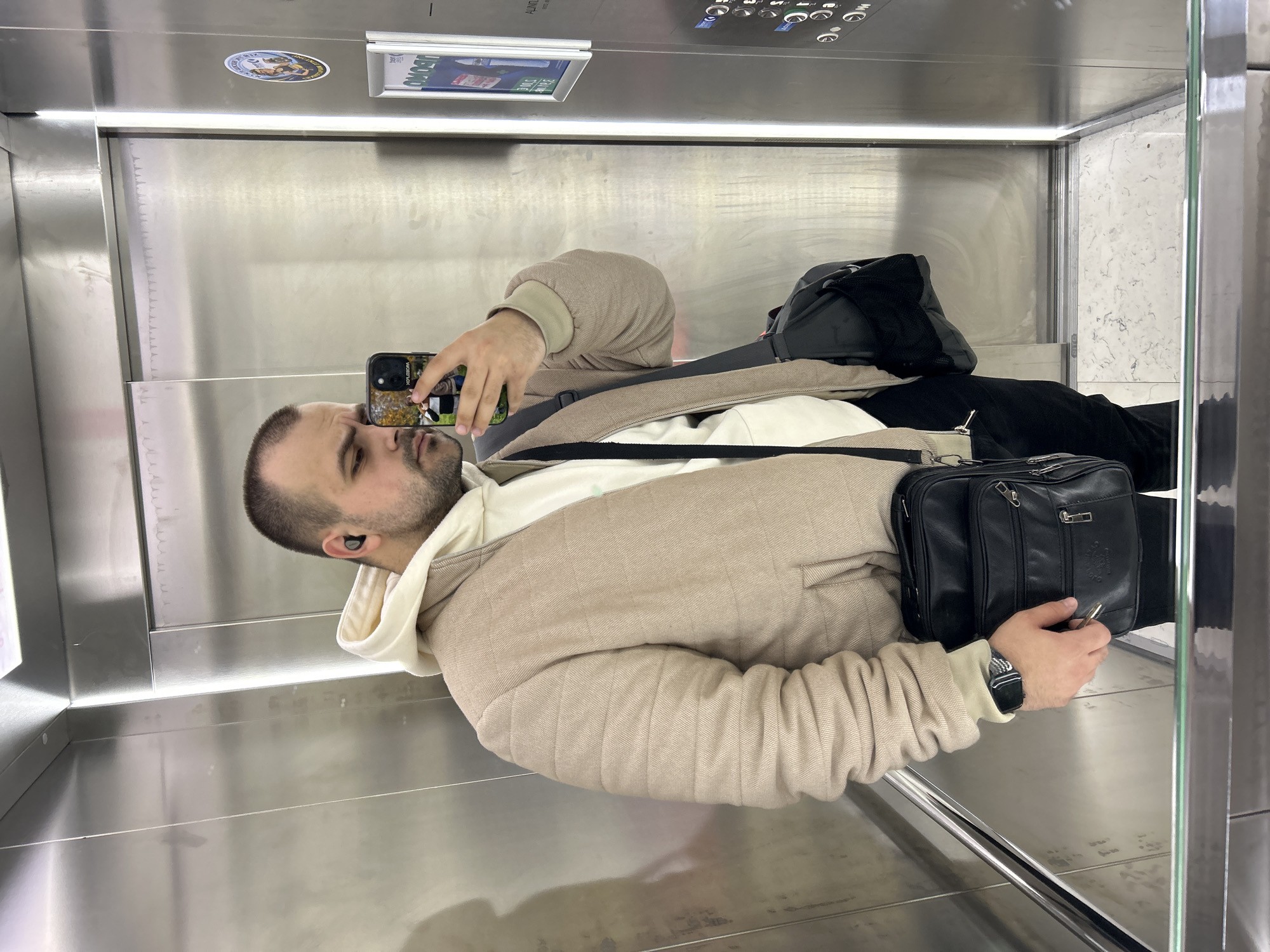 A man is taking a photo of himself in an elevator. He has short brown hair and a beard. He is wearing a beige coat over a white hoodie. He is holding a smartphone in his left hand. The elevator has stainless steel walls. There are two signs visible on the top of the wall, but they are out of focus and illegible. The location is unknown and not specified in the information provided.