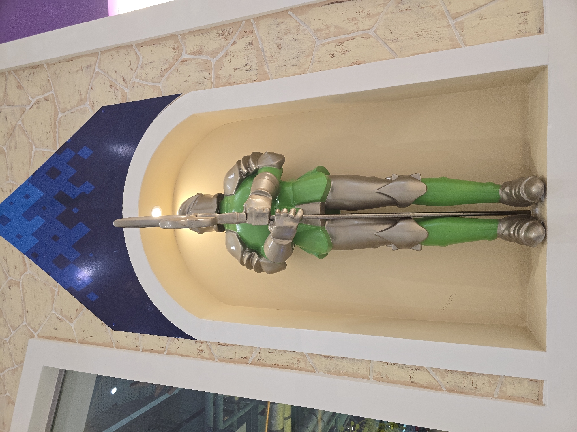 A horizontal eye-level shot depicts a statue of a green and silver knight, lying down, holding a lance in a semi-circular, recessed niche in a wall in Grozny, Russia.
The knight is predominantly green with silver colored armor parts. He lies flat on his back within the niche. He is holding a silver-colored lance with both hands across his chest.
The niche is a semicircular shape with a light yellow interior. It is bordered by a white trim and a stone-like exterior. To the left of the niche, there is a large blue area, with blue patterns, that forms a downward pointing triangle.
Below the niche is a window. Above the niche is a faux brick pattern. At the top left, there is a purple wall.