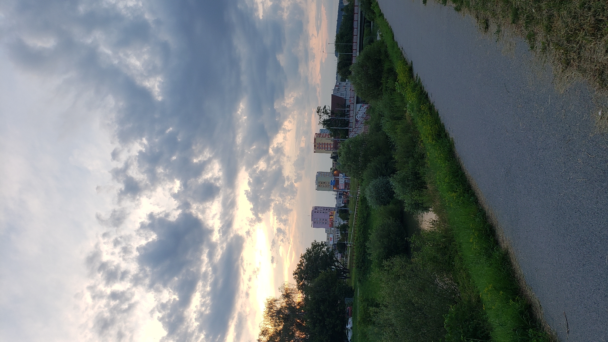 The image captures a scenic view of the city of Prešov, Slovakia, during a cloudy sunset. In the foreground, a paved path runs diagonally across the frame, bordered by lush green grass and vegetation. To the left of the path, a river flows gently, flanked by dense trees and bushes. In the background, the city skyline is dotted with several tall buildings, including apartment blocks with colorful facades. The sky above is a dramatic canvas of grey and white clouds, with hints of warm sunlight breaking through, creating a serene and picturesque atmosphere. The overall scene suggests a peaceful evening in a city park or along a riverbank, offering a glimpse of urban life harmoniously blended with nature.
