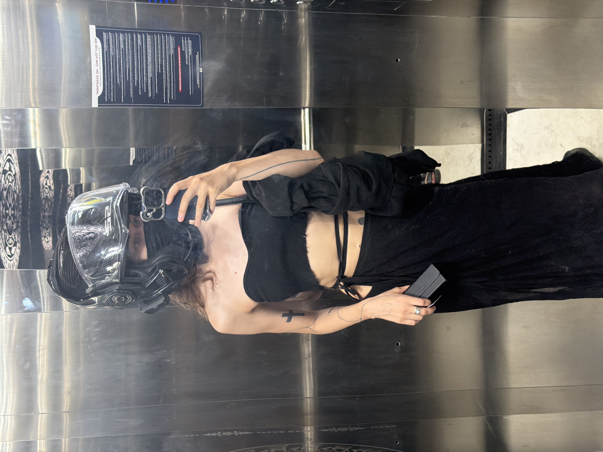 The image depicts a person wearing a futuristic helmet and a black outfit, possibly in an elevator. The elevator has a stainless steel interior, featuring a safety instruction placard. The person is holding a smartphone, suggesting they might be taking a selfie or recording a video. The location is unknown.