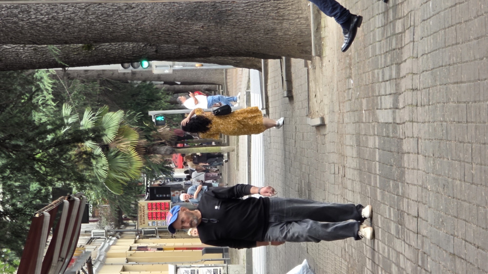 A man wearing a black long-sleeved shirt, grey jeans, and a blue baseball cap walks forward on a brick-paved sidewalk. He is in the foreground, slightly to the left of the frame. Behind him, a woman in a yellow floral dress walks away from the camera, also on the sidewalk. Further in the background, several other people are walking and standing. A traffic light with a green signal is visible to the right of the frame. A sign with currency exchange rates is displayed on a building on the left side of the image. The scene appears to be a street in the city of Batumi, Georgia, as indicated by the currency exchange sign which shows rates for USD, EUR, RUB, and TRY. The environment includes trees lining the street and buildings in the background.