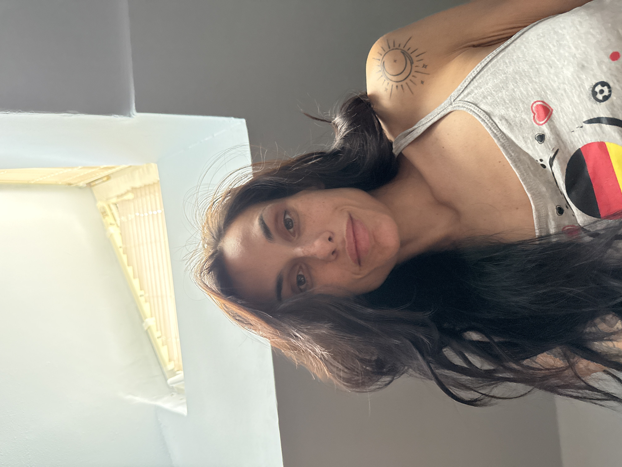 A portrait-style image, likely a selfie, captures a woman with long, dark brown hair looking directly at the camera with a subtle, neutral expression. The photograph is oriented sideways, with the woman's head tilted. She has brown eyes and a fair complexion.
She is wearing a light gray tank top adorned with various colorful motifs, including a red heart, a black and white soccer ball, and a circular design featuring horizontal stripes of black, yellow, and red. A prominent tattoo of a stylized sun encircling a crescent moon with small stars is visible on her left shoulder.
The setting appears to be an indoor room, likely in Pleven, Bulgaria. Bright natural light illuminates the scene, coming from a window or skylight positioned to the upper left of the frame (viewer's left). This window has a light-colored frame, possibly made of wooden or bamboo slats, suggesting a daytime environment. The walls of the room are a light, neutral color, contributing to the bright atmosphere.