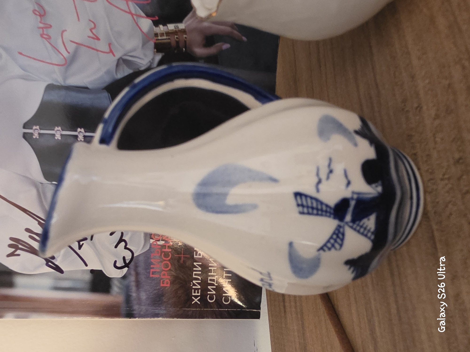 The image shows a white ceramic pitcher with blue windmill and floral decorations. The pitcher is in the foreground, resting on a wooden surface. In the background, a person is visible, wearing a white shirt with pink writing on it. Part of a magazine or book cover is also visible, with text in Cyrillic alphabet, likely in Russian or Bulgarian. The person's hand is reaching towards the pitcher. The scene appears to be indoors, with natural lighting. The overall impression is a still life arrangement, possibly showcasing a decorative object. The visible text on the magazine is "ПИРА БРОСЧЕ ХЕЙЛИ Б СИДНИ Б СИЧТ". There is also a watermark "Galaxy S26 Ultra" on the right side of the image.