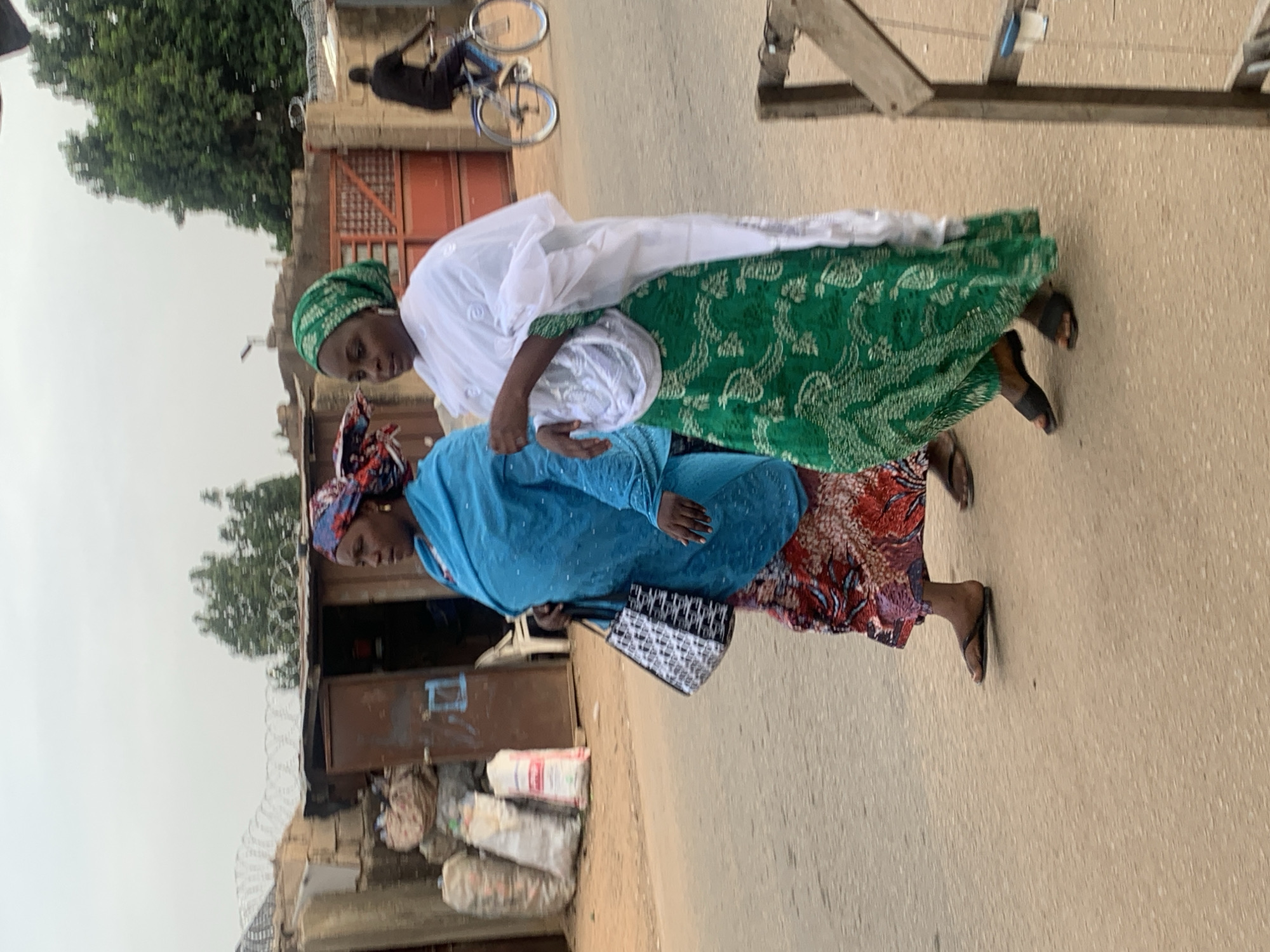 Two women are walking on a paved road. The woman in front is wearing a green and white patterned robe and head covering. The second woman is wearing a blue robe, a patterned head covering, and carrying a purse. They are walking past a building and some sacks that are stacked next to it. The image appears to be in Jos, Nigeria. In the background, a man is riding a bicycle near a gate.