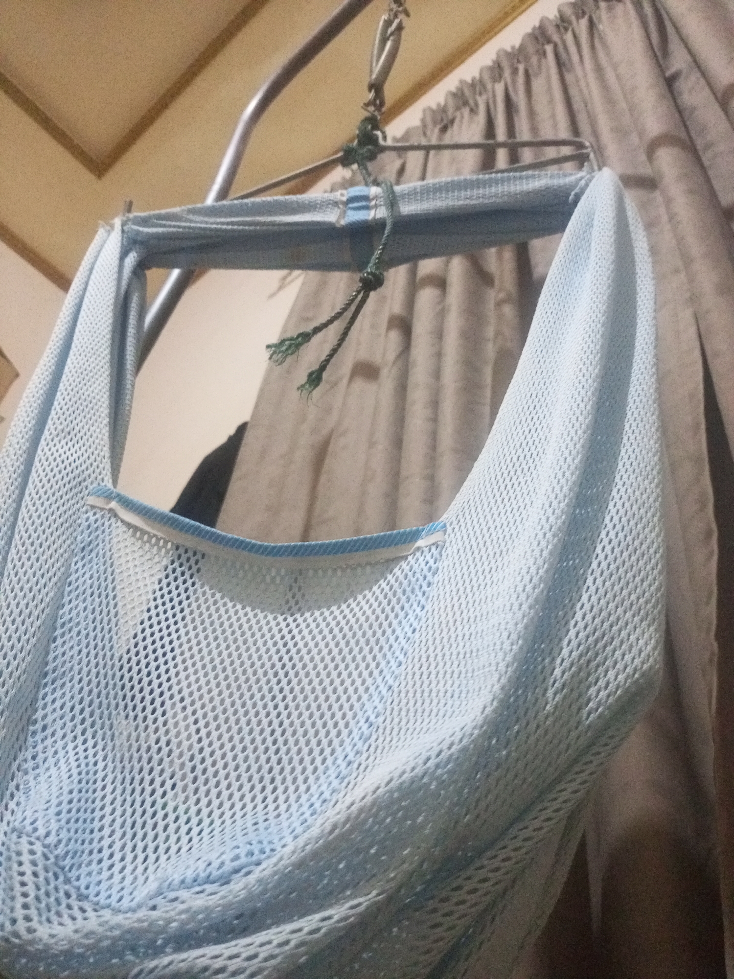 A close-up, low-angle shot shows a light blue, mesh-like fabric draped over a metal bar. The fabric appears to be part of a hanging hammock or sling, with a white and blue trim visible along its upper edge. Green ropes, frayed at the ends, are tied to the metal bar, supporting the fabric. In the background, pleated beige curtains hang behind the metal bar, and a light-colored ceiling with visible seams is above. The scene is dimly lit, with shadows cast on the fabric and a dark, indistinct shape visible behind the mesh. The image does not contain enough information to determine its location in Lhokseumawe, Indonesia.