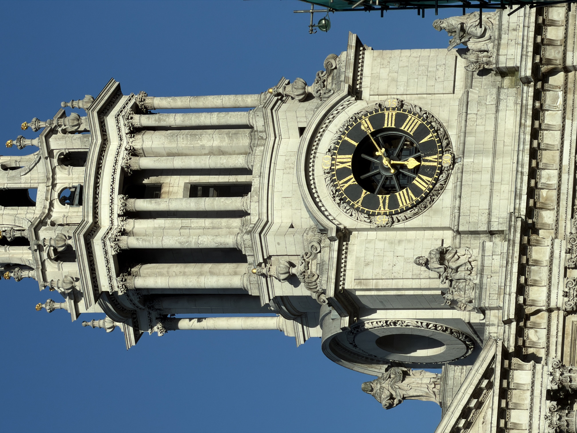 This vertical close-up photograph captures the ornate architectural details of a grand clock tower, set against a vibrant, cloudless blue sky. The structure is built from light-colored stone, likely limestone, and features an abundance of intricate carvings, columns, and decorative elements.
The focal point is a large, circular clock face, predominantly black with striking gold Roman numerals. The golden minute and hour hands clearly indicate the time as approximately 10:45. Surrounding the clock face is an elaborate stone border with detailed floral or scrollwork patterns.
Above and to the left of the clock, the tower rises with multiple tiers of fluted or plain columns supporting decorative cornices and balustrades. Ornate stone sculptures, some appearing as gargoyles or stylized figures, adorn various ledges and corners of the architecture. Towards the top, golden finials add a touch of regality to the spires. Scaffolding and green netting are visible at the very top edge of the image, suggesting ongoing maintenance or construction work on the upper sections of the building. The bright, direct sunlight highlights the textures and shadows of the stonework, emphasizing the depth and craftsmanship. Given the context, this is a historic building in London, United Kingdom.