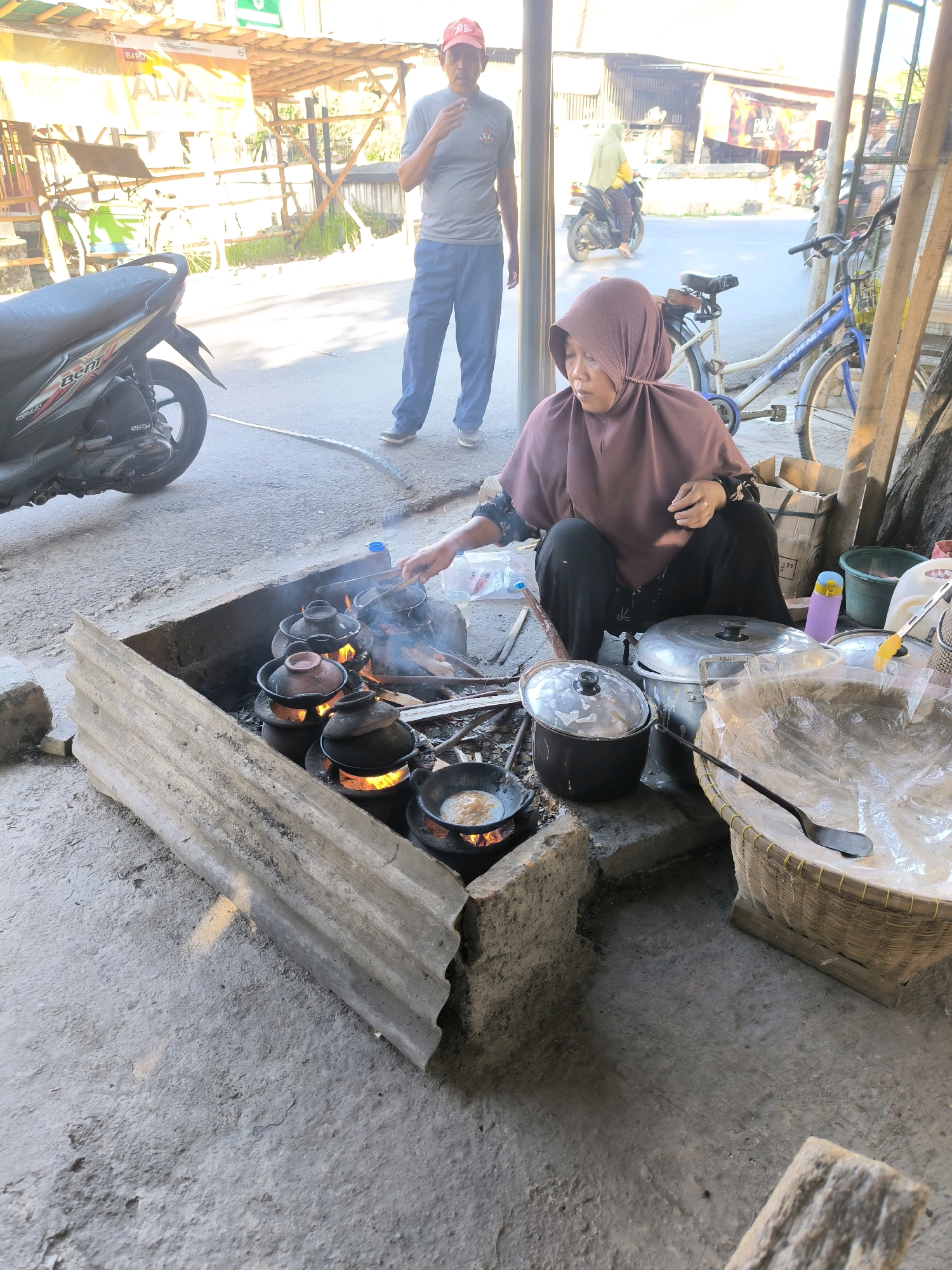 A woman wearing a mauve hijab and black pants kneels in front of a cooking fire in Karanganyar, Indonesia, stirring small clay pots with a spatula. A man in a grey t-shirt and blue pants stands behind her, holding a cigarette. The scene appears to be an outdoor street food stall, with a motorcycle parked to the left and a bicycle to the right. Various cooking utensils, including large pots and a wok, are arranged around the fire. The background suggests a bustling urban environment with buildings and other stalls. The ground is paved with concrete.