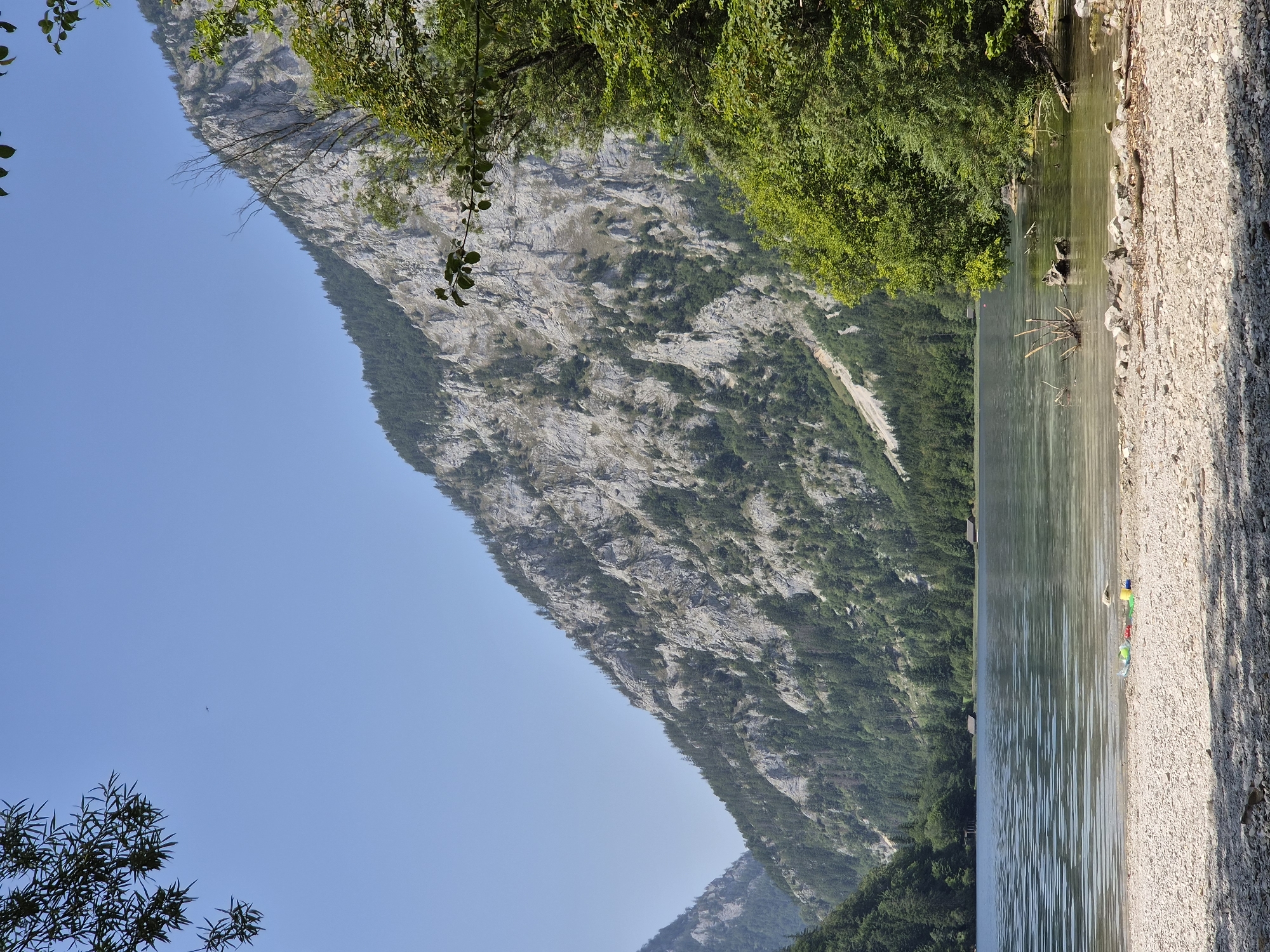 This is a vertical, eye-level, medium shot of a serene lake scene on a clear, sunny day. The foreground is a pebbly beach with several colorful children's toys scattered near the water's edge, including a red bucket, a yellow shovel, and some green plastic items. A few pieces of driftwood are partially submerged in the shallow water.
The lake itself is calm, with gentle ripples reflecting the sky and surrounding landscape. Small, dark wooden structures, possibly boathouses or cabins, are visible along the far shore, nestled amongst a dense forest of green trees.
Dominating the background are imposing, steep mountains with rocky, grey faces, partially covered by the same lush green forest that lines the lake's edge. The sky above is a clear, pale blue. The overall impression is one of natural beauty and tranquility. The image appears to be of the Präbichl lake, near Eisenerz, Austria, a popular recreational area known for its scenic mountain vistas and lake activities.