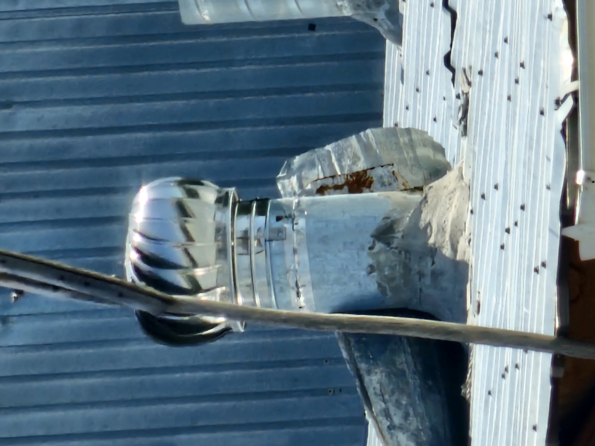 This is a medium-shot, eye-level, outdoor photograph taken on a sunny day. The scene depicts a rooftop with a corrugated metal finish, showing some signs of wear and tear, including patches of rust and loose material. In the foreground, a vertical wooden pole is positioned slightly off-center to the left. To the right of the pole is a metal roof vent, specifically a turbine vent, made of shiny, reflective metal with rotating blades. The vent is mounted on a cylindrical metal pipe, which appears to be connected to the roof. The base of the vent and pipe show signs of damage and weathering, with some sections appearing to be broken or corroded. Behind the vent is a blue corrugated metal wall, likely part of a building. The overall environment suggests an industrial or utility area, possibly in a less developed part of the city of Özgön, Kyrgyzstan, given the state of disrepair of some of the structures. There are no people or animals visible in the frame.