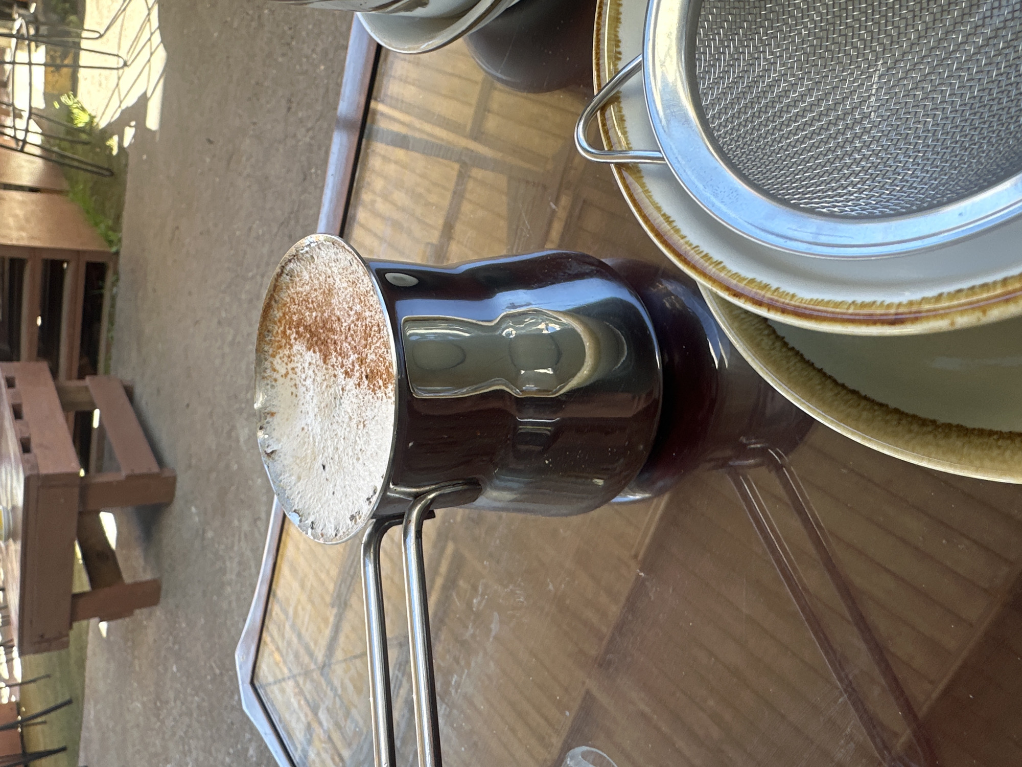 The image shows a close-up of a coffee cezve filled with frothy coffee, dusted with cinnamon, resting on a glossy, dark brown table. The cezve is made of stainless steel with two handles. In the foreground, a stack of plates and a fine-mesh sieve are visible. The background suggests an outdoor setting, possibly a cafe, with a wooden bench and some greenery, hinting at a location in Bellingen, Australia. The lighting indicates it is daytime, with sunlight casting shadows.