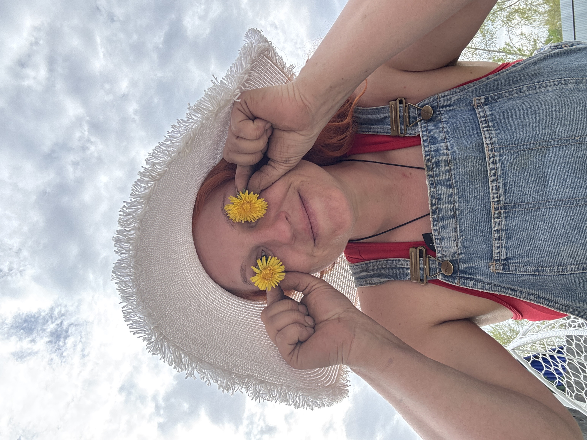 A woman with red hair, wearing a white straw hat, a red tank top, and denim overalls, is holding two yellow dandelions over her eyes. The photo appears to have been taken from a low angle, looking up at the woman and the sky. The sky is cloudy. It's not possible to confirm the location of Chaikovsky, Russia from the visual data.