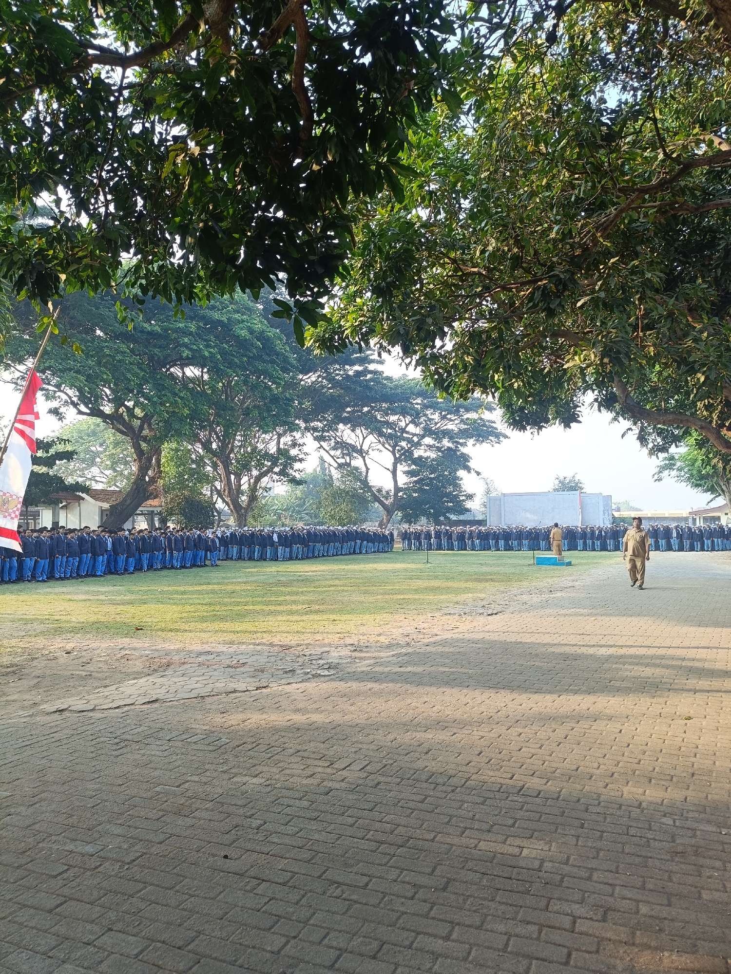 In Kediri City, Indonesia, this image captures an outdoor assembly on a paved area bordering a grassy field, bathed in early morning light filtering through dense trees. A long line of young people, presumably students, dressed in identical navy blue uniforms and blue trousers, stand rigidly in formation on the grass. A red and white Indonesian flag, partially unfurled and bearing a crest, is positioned on the left side of the frame, attached to a flagpole. In the mid-ground, a few individuals in khaki uniforms are visible near a small blue podium. One person in khaki walks towards the right foreground, casting a long shadow on the brick-patterned pavement. Behind the students, a long, low building with a white facade and a flat roof is visible, further back are several large, mature trees, their branches arching over the scene. The overall atmosphere suggests a formal gathering, possibly a school assembly or a ceremony, taking place in a well-maintained outdoor space.
