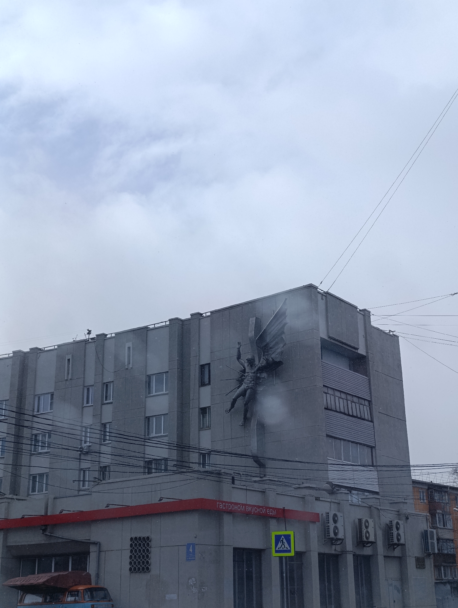 The image shows a large, gray building in what appears to be urban Krasnoyarsk, Russia. The building has several stories with visible windows. On the corner of the building is a high-relief sculpture depicting a winged figure, possibly an angel or mythical being. A smaller building or extension is visible at the base of the larger structure, featuring a red trim and a sign written in Russian, "ГАСТРОНОМ ВКУСНОЙ ЕДЫ," which translates to "Gastronom of Delicious Food". Below the sign are several air conditioning units. A pedestrian crossing sign is visible. Overhead, numerous power lines crisscross the view. The sky is overcast, and there appear to be light snow or precipitation falling. There is also a blue van in the lower left corner of the image.