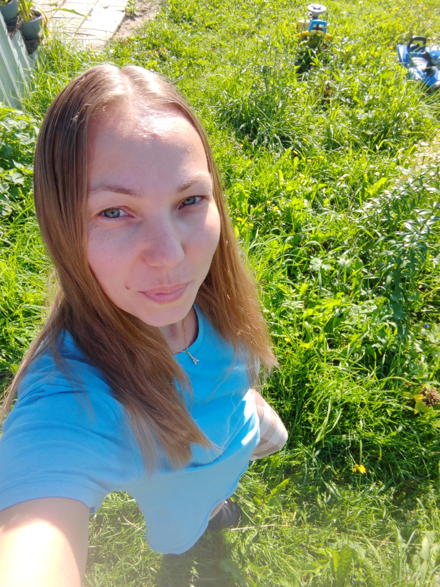 A young woman with blonde hair is taking a selfie outdoors. She is wearing a blue t-shirt and a necklace. She is standing in a grassy area with some toys in the background. The lighting suggests it is daytime, and the environment appears to be a park or backyard.