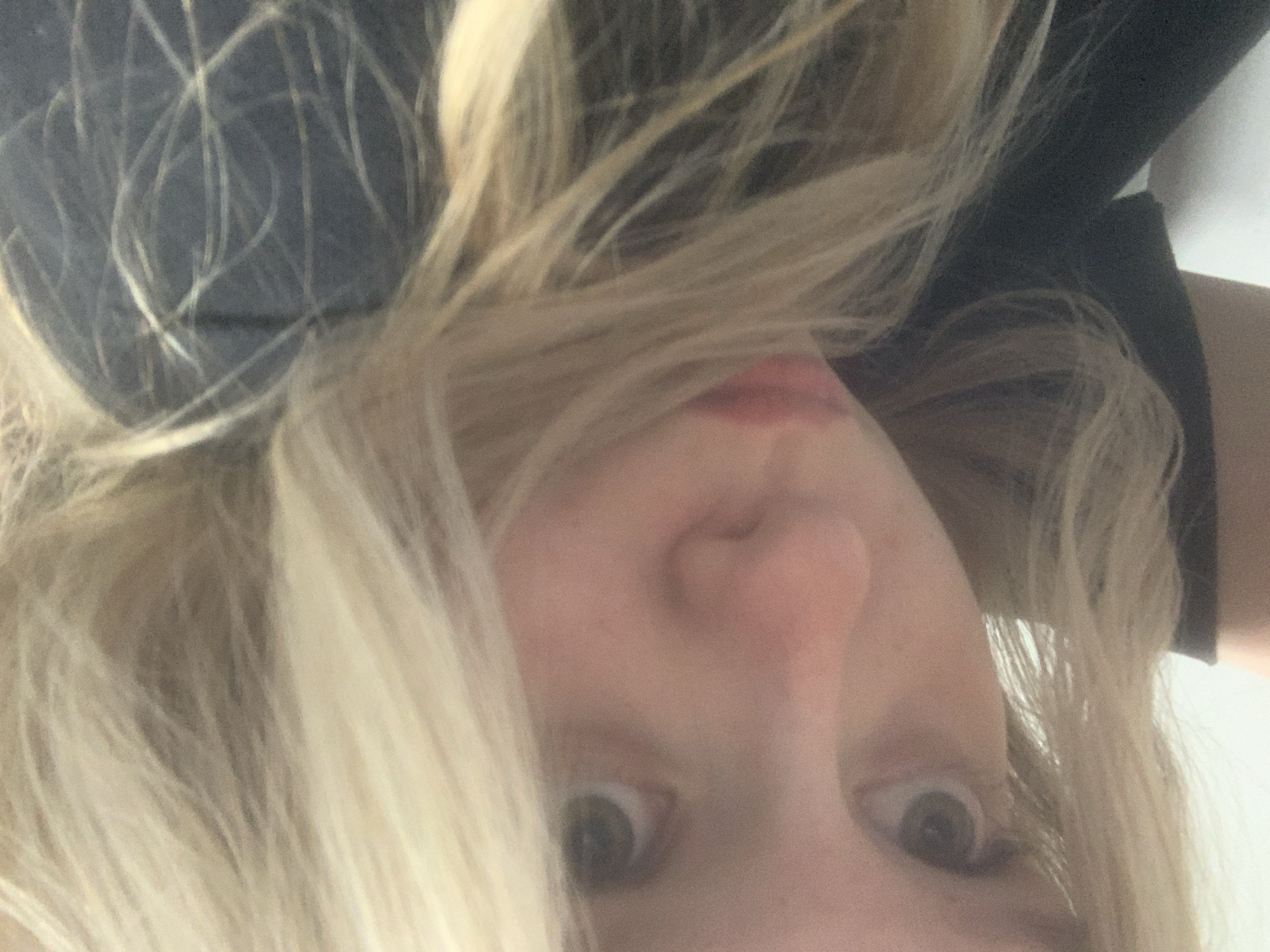 This is a close-up, vertically oriented selfie of a young person with blonde hair. The person is looking directly at the camera, with a slight open-mouthed expression. The setting appears to be indoors, with a plain white wall visible in the background on the right side. The person is wearing a dark-colored garment, possibly a shirt or a jacket. The lighting is bright, suggesting daylight or well-lit indoor conditions. The image is slightly blurry, indicating movement or a focus issue. No text is visible in the image. The scene is intimate and personal, capturing a moment of direct engagement with the viewer.