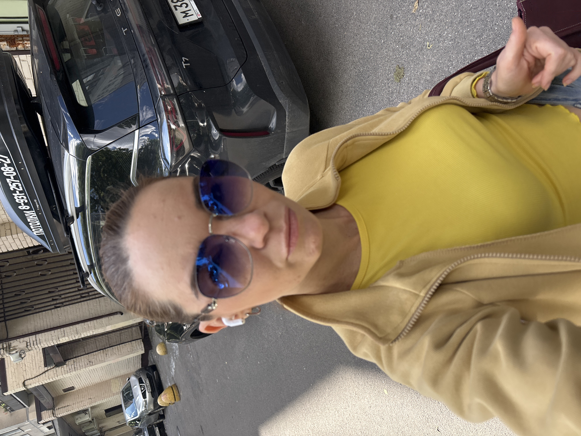 The image shows a woman with brown hair, wearing blue-tinted sunglasses and a yellow top, with a tan jacket zipped up partially. She has white earbuds in her ears and appears to be taking a selfie. She is outdoors in what appears to be a city street with buildings on the left and parked cars behind her. One car is black and has a roof box with Cyrillic writing. Another car, silver, is parked further down the street. The environment suggests a residential or commercial area within a city.