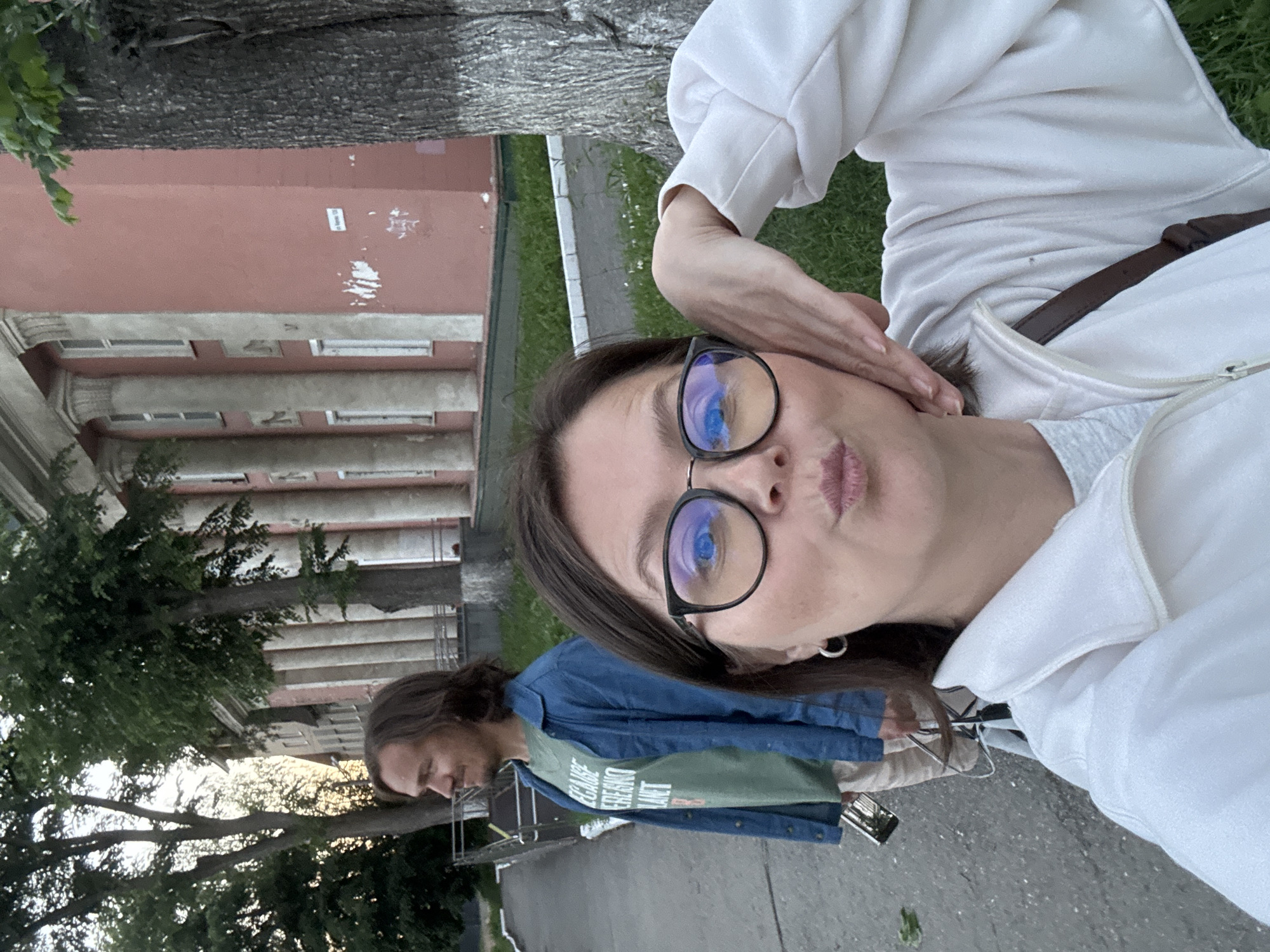A medium shot captures a woman in the foreground, looking directly at the camera with her lips pursed and her right hand resting on her cheek. She is wearing a white zip-up jacket over a grey t-shirt and large black-rimmed glasses. A dark brown strap of a bag is visible over her shoulder. In the background, a man with long brown hair and a blue denim jacket over a green t-shirt stands looking to his left. He is holding a metallic object in his left hand. The two are positioned on a paved walkway next to a grassy area. Behind them, a pink building with large columns and several windows is visible, partially obscured by trees. The scene appears to be outdoors, possibly in a park or near a public building in the city of Izhevsk, Russia, given the architectural style.
