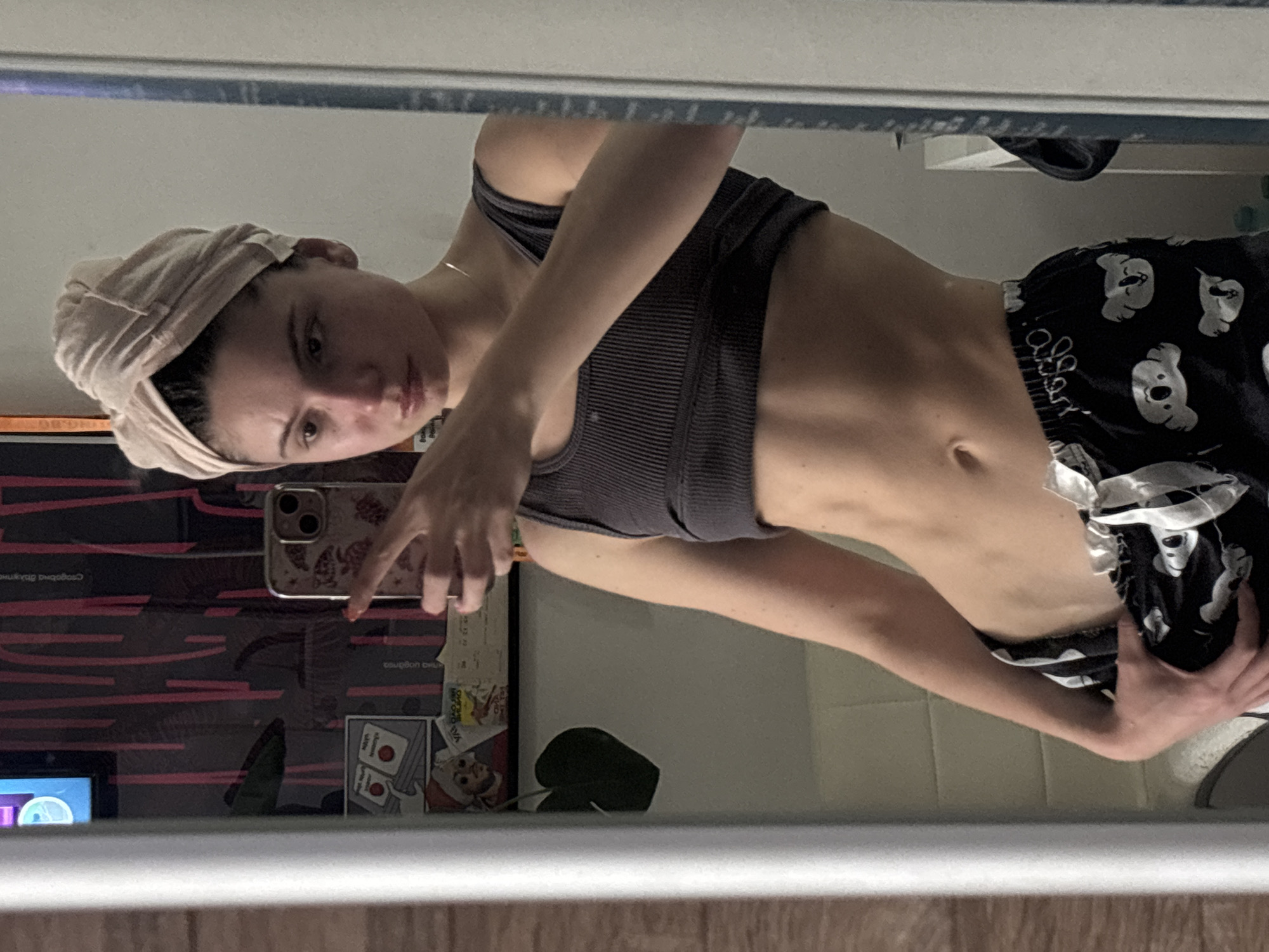 A woman is posing for a selfie in front of a mirror. She is wearing a dark grey sports bra and black pajama shorts with white koala bear prints. A light pink towel is wrapped around her head. She is holding a cell phone with a floral patterned case. The reflection shows a television screen with colorful graphics and some stickers and cards attached to the side. The environment appears to be a bathroom or bedroom. The location is Unknown, Unknown.