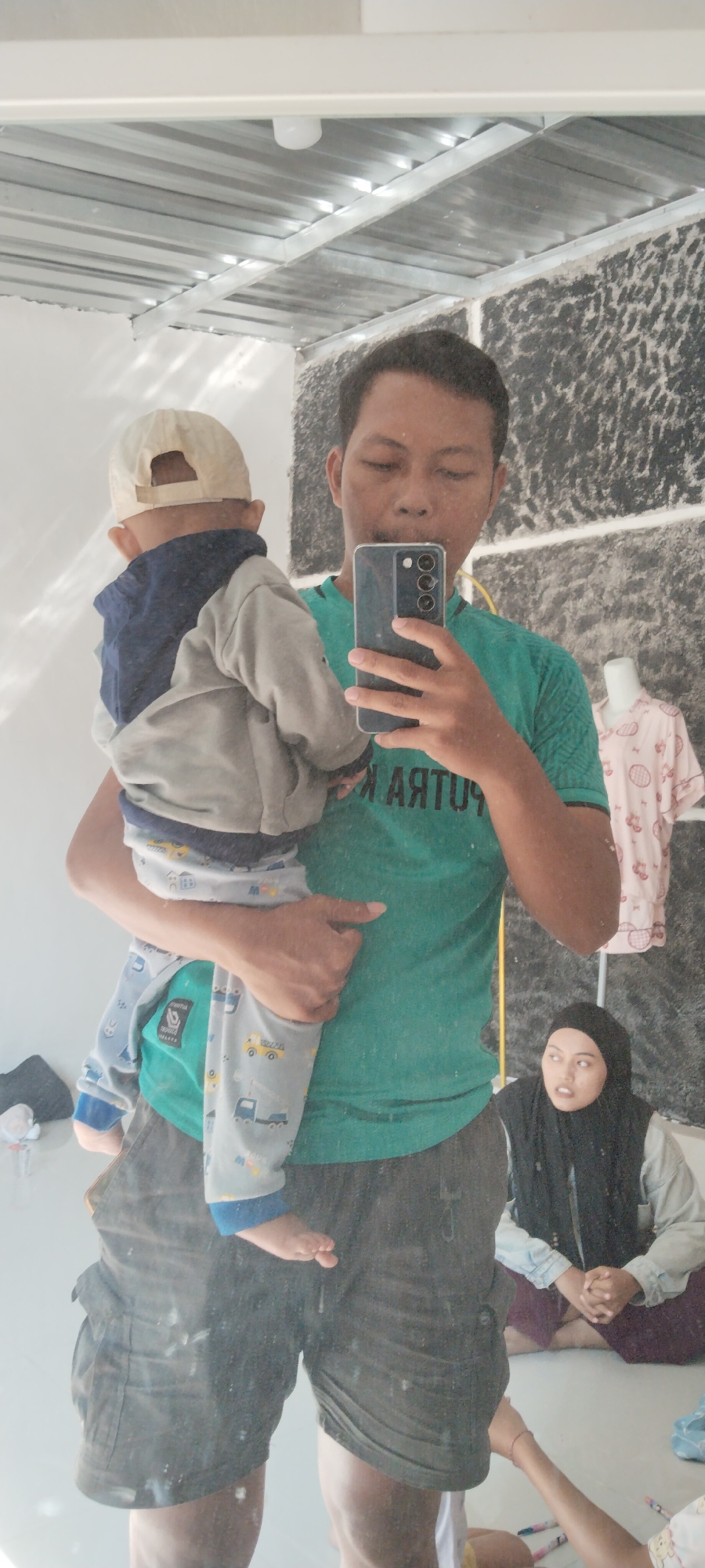 In a brightly lit indoor space, likely a room in Kecamatan Pati, Indonesia, a man is taking a mirror selfie while holding a young child. The man, wearing a green t-shirt and dark shorts, is positioned in the center of the frame, holding the child who is facing away from the camera and wearing a beige cap and a grey jacket over a blue top. The child's legs are clad in light grey pants with a pattern of yellow and blue trucks.
In the reflection of the mirror, another woman is visible sitting on the floor to the right of the man, wearing a black hijab and a light-colored outer garment over a dark purple skirt. Her hands are clasped in her lap, and she is looking towards the man and child. A white mannequin wearing a pink patterned shirt is also visible in the background to the right.
The environment appears to be under construction or in a state of disarray, with exposed ceiling beams and a textured, dark-colored wall behind the subjects. The floor is light-colored and shows some smudges or dirt. The lighting is harsh, casting reflections and shadows within the mirror. A yellow pole is visible in the reflection, extending from floor to ceiling.