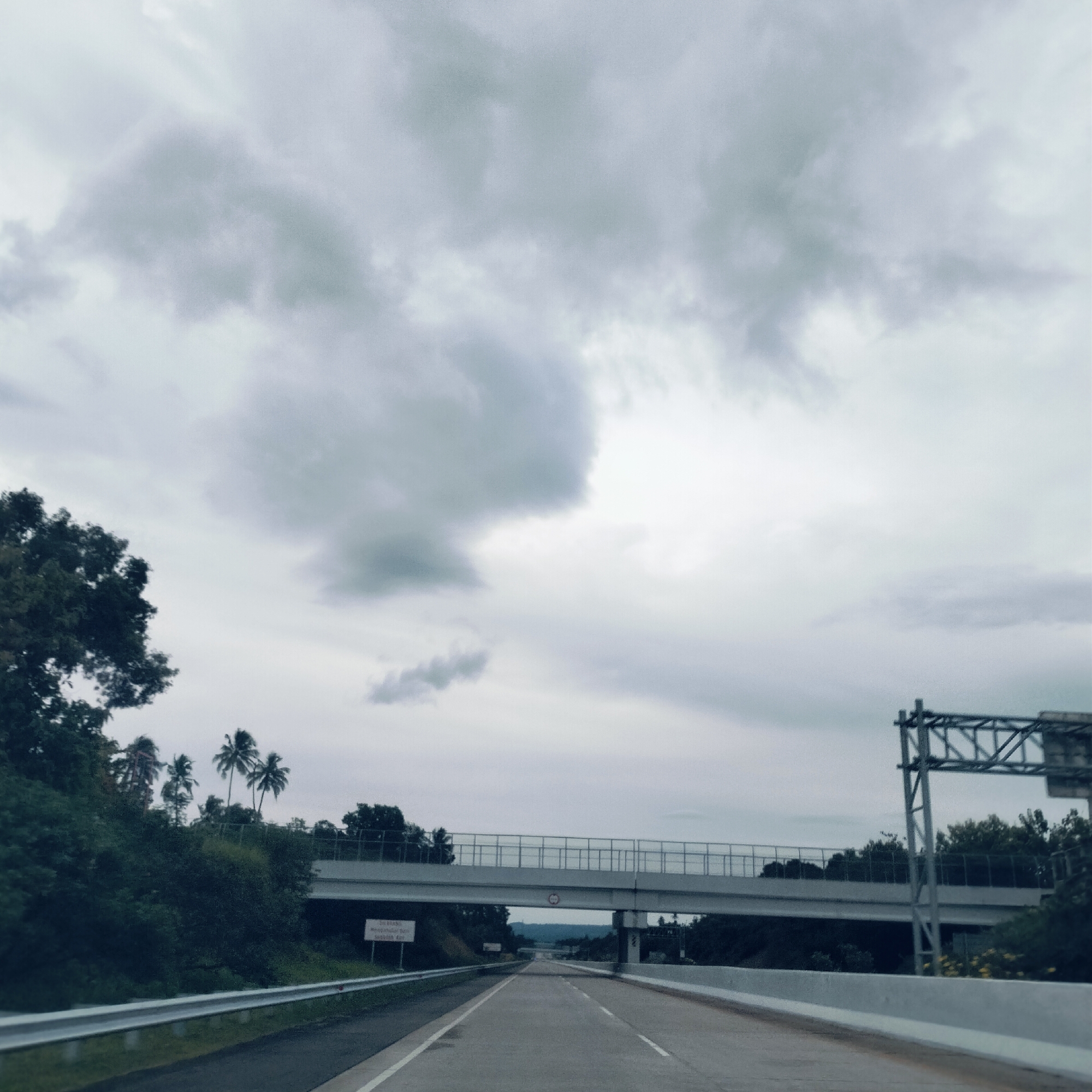 The image captures a wide-angle view of a highway under an overcast sky in Lampanahtunong, Indonesia. The perspective is from inside a vehicle, looking straight down a multi-lane asphalt road that stretches into the distance.
The sky is dominated by thick, grey clouds, indicating a heavily overcast day with muted lighting. Lush green vegetation, including trees and several palm trees, flanks both sides of the highway, particularly dense on the left.
In the midground, a concrete overpass or pedestrian bridge spans the highway. Underneath this overpass, on the left side of the road, a white rectangular sign with text in an Indonesian language is visible but mostly illegible due to blur and distance. A red circular sign with "4.2m" inside, indicating a height restriction, is affixed to the underside of the overpass.
On the right side of the road, an overhead gantry structure, likely for road signs or surveillance equipment, stands tall. Guardrails line both sides of the highway, and concrete barriers are also visible. The road itself appears clear of traffic ahead, suggesting a relatively quiet period or remote location. The overall atmosphere is calm and subdued due to the cloudy weather.