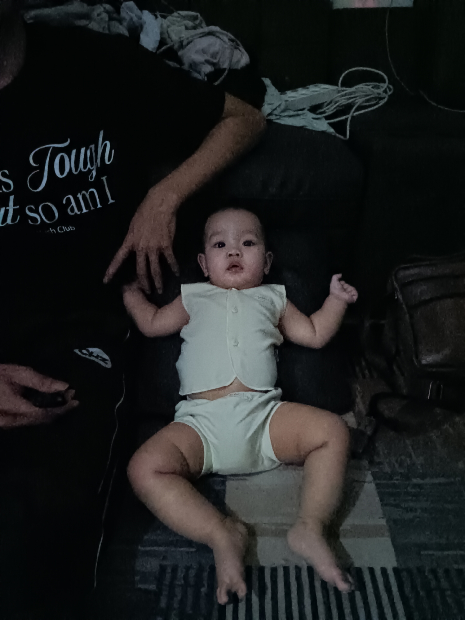 An infant lies on a patterned rug next to an adult who is partially visible, wearing a black t-shirt with white text that reads "Tough so am I". The infant is wearing a light-colored two-piece outfit, consisting of a sleeveless vest and shorts. The adult's left arm rests on the infant's legs, and their right hand is extended towards the infant. A dark-colored bag is visible to the right of the infant. The environment appears to be an indoor setting, possibly a living room, with a dark-colored sofa or couch behind the subjects. The lighting is dim, creating shadows and a slightly grainy image quality.