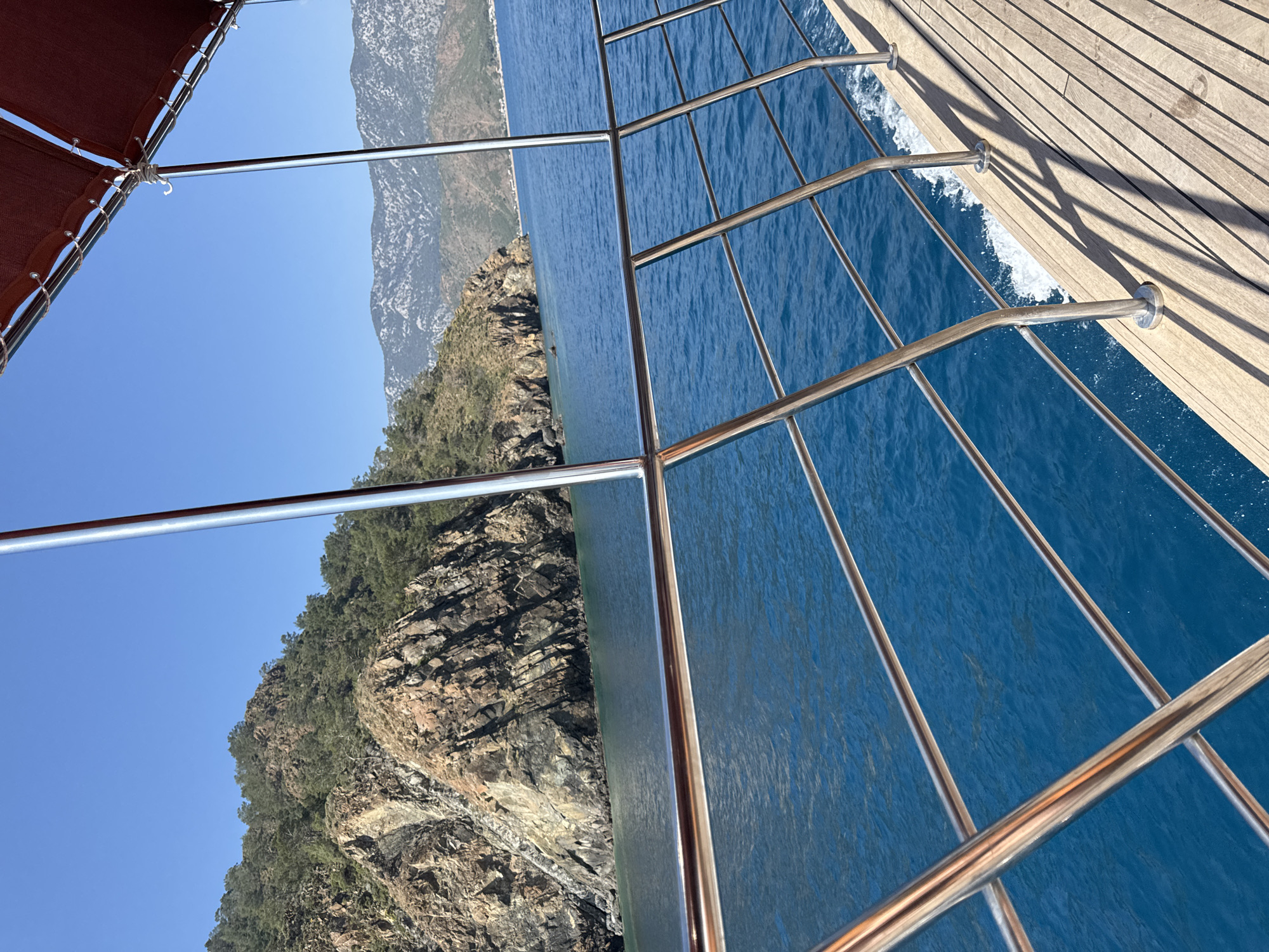 The image captures a vibrant daytime scene from the deck of a boat cruising along a mountainous coastline, likely in Kumluca, Turkey. The perspective looks out over the water towards the land.
In the foreground, a section of the boat's wooden deck, featuring light-colored planks, is visible on the right. Chrome railings, part of the boat's perimeter, extend across the lower half and right side of the frame, creating a grid pattern. White foamy water, churned by the boat's movement, can be seen just below the deck, sparkling against the deep blue sea.
The sea, a brilliant cerulean blue, dominates the central and right-hand portions of the image, showing gentle ripples and reflections. It stretches towards a rugged, rocky coastline that rises steeply from the water on the left side of the frame. The mountains display prominent grey and brown rock faces, interspersed with areas of lush green vegetation, including trees and bushes. The coastline extends into the distance, appearing softer and hazier under the bright light.
Above the land and sea, the sky is a clear, cloudless blue, suggesting excellent weather conditions. In the upper left corner, a small part of a dark red or maroon canvas canopy, possibly a sunshade, is visible, held by ropes and metal supports, providing a hint of the boat's structure overhead.
The scene conveys a sense of tranquility and leisure, consistent with a boat trip or cruise. The bright, direct sunlight indicates it is likely mid-day or early afternoon. No visible text is present in the image.