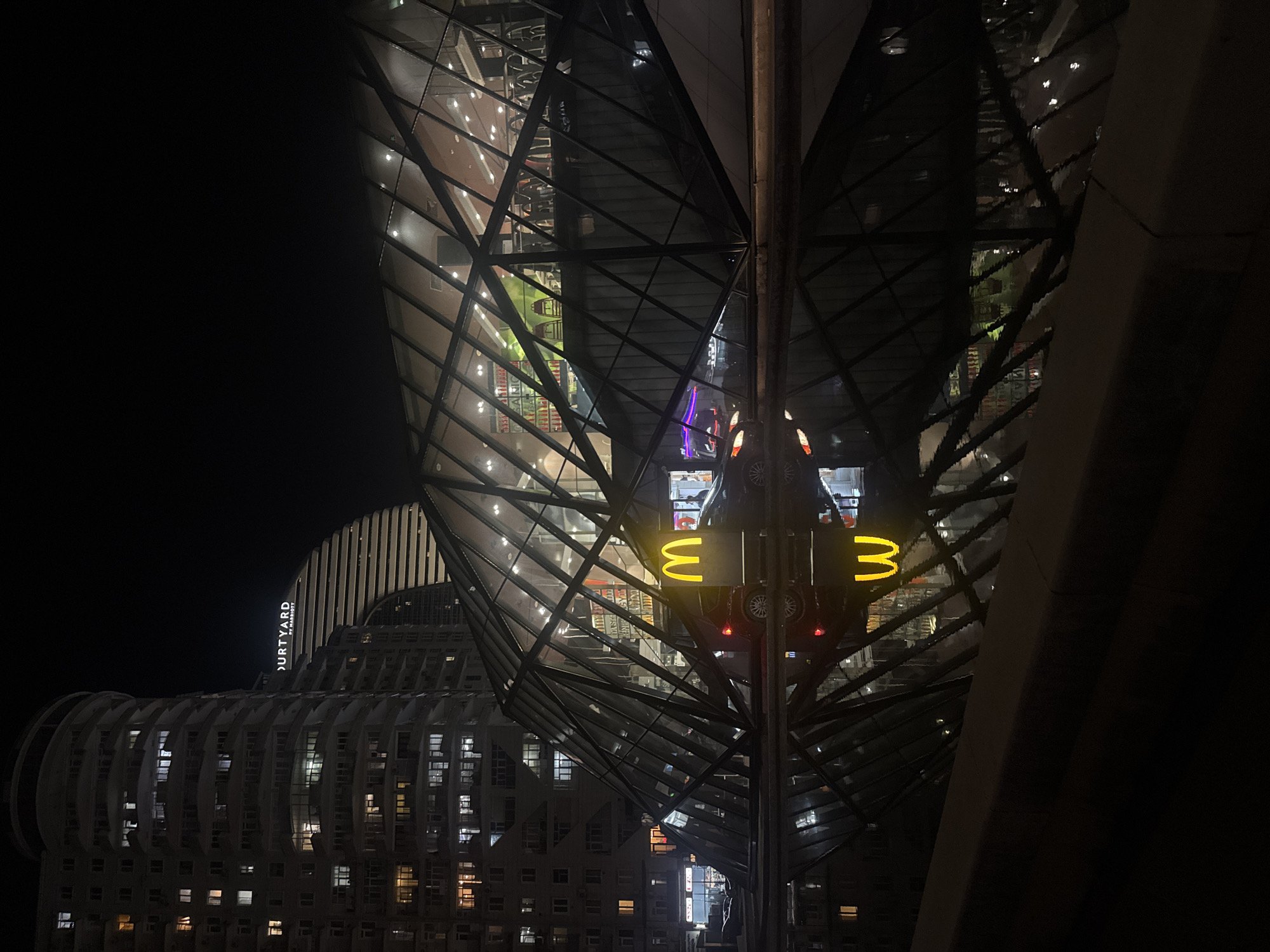 The image shows a night view of a modern building, with the text "Courtyard by Marriott" vertically visible on the left side of the frame. To the right, there is a spiral-shaped structure constructed of glass and metal. A vertical pole with three upside-down "M" shapes is visible in the center, with two tail-lights below. The background is primarily dark.