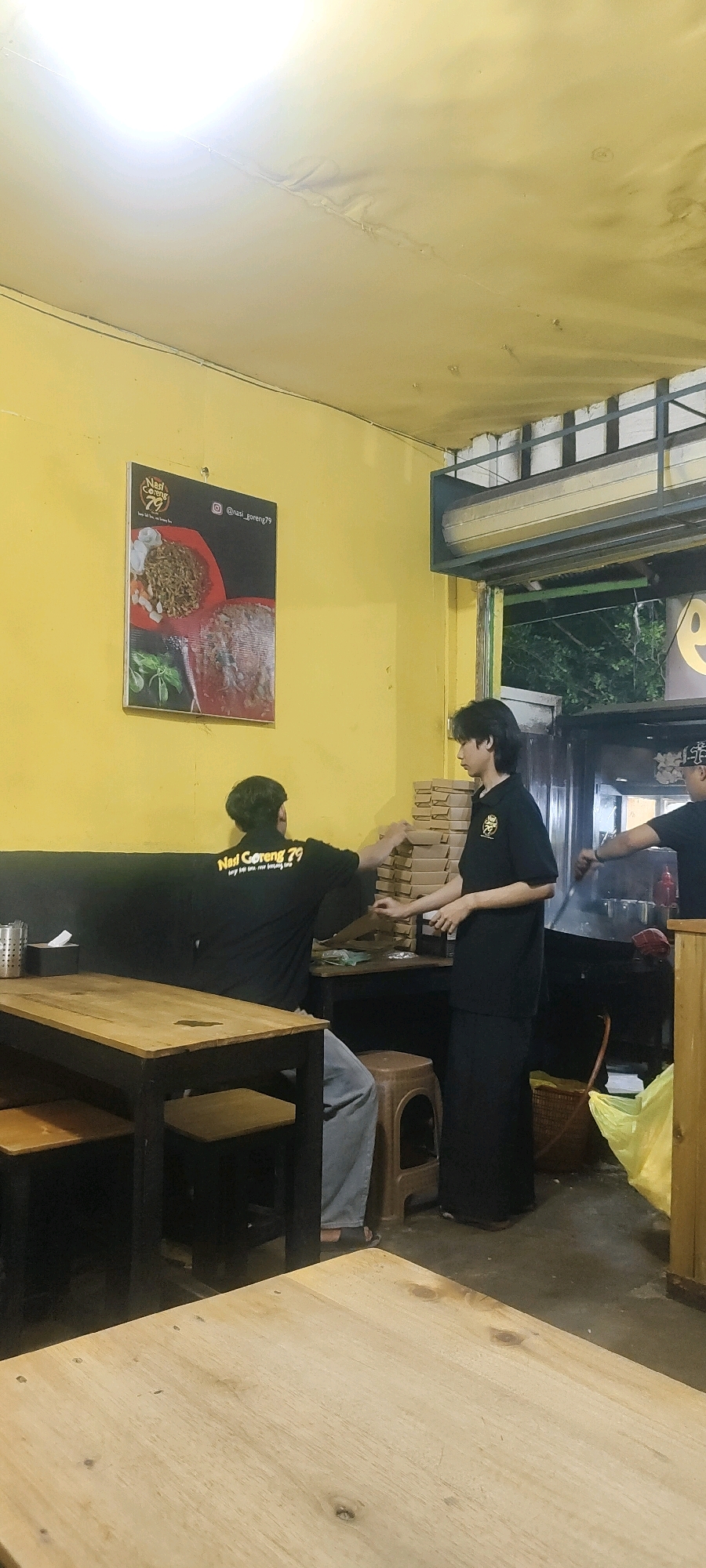 In the city of Palangka Raya, Indonesia, within an indoor establishment, three individuals are present. One individual, wearing a black t-shirt with text that appears to say "Nasi Goreng 79", is seated facing away from the camera and is engaged in handling items on a table. Another individual, also wearing a black t-shirt, stands to the right of the first, facing them and holding what looks like stacks of pizza boxes. A third individual, partially visible on the far right, wears a black t-shirt and is near a cooking area. The environment features yellow walls, wooden tables and chairs, and a poster on the wall depicting food. The lighting suggests it is evening or indoors.