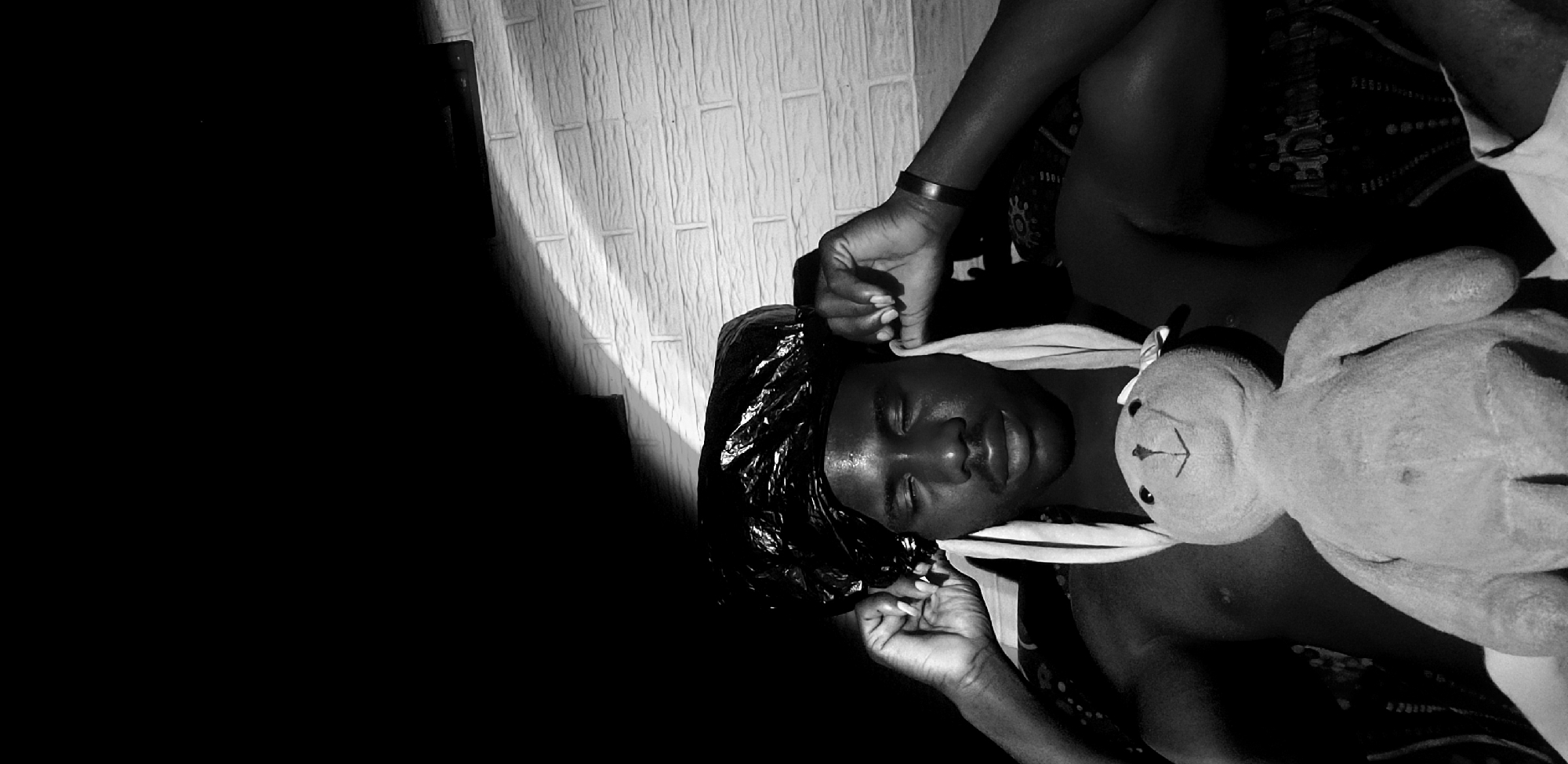 A black and white, medium shot photograph shows a Black person, appearing to be an adult male, with his eyes closed and a black plastic bag covering his head. The person is holding the edges of the bag with his hands, near his ears. Two white fabric strips hang down from the bag, resembling rabbit ears. A plush toy, resembling a rabbit with a sewn-on smile and two small ears, is held in front of the person's chest. The person is shirtless, and a dark-colored bracelet is visible on his left wrist. The background consists of a dark wall on the left, transitioning to a textured white brick wall on the right, with a diagonal shadow cast across it from the left. The overall scene appears to be indoors. The provided information states this image was taken in Narayi, Nigeria.