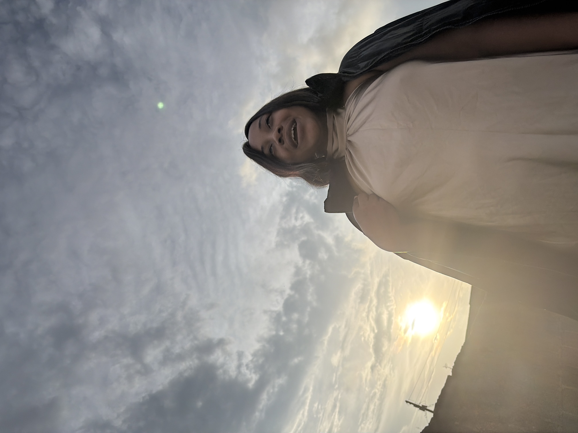 The image is a low-angle shot of a young Black woman standing outside, looking up and to her right with a smile. She has dark hair and is wearing a light-colored halter top and a dark jacket draped over her shoulders. The background consists of a cloudy sky with the sun setting or rising in the lower left corner, casting a warm glow and creating lens flare. The sun's light illuminates part of a low-rise structure or wall in the foreground to the left, with what appears to be utility poles and wires visible against the bright sky. The scene suggests an outdoor setting in the city of Jos, Nigeria, possibly during an evening or early morning.