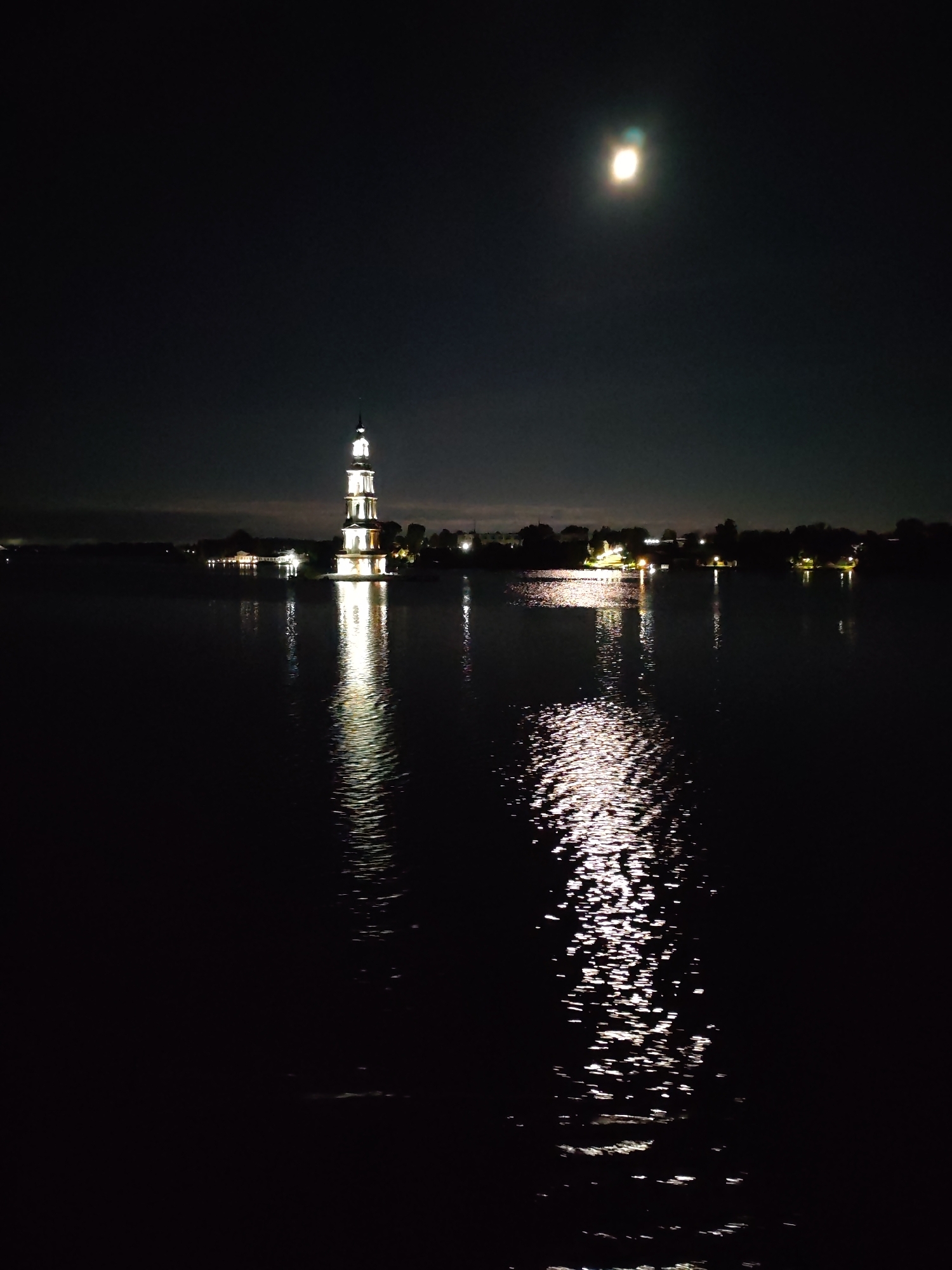 The image depicts the bell tower of Saint Nicholas Monastery in Kalyazin, Russia, illuminated at night. The tall, multi-tiered structure stands in the water, with its lights reflecting brightly on the dark surface. In the upper right portion of the frame, the moon is visible as a bright, circular object in the night sky, also casting a reflection on the water. The surrounding shoreline is dark, with faint outlines of buildings and trees visible in the distance. The overall atmosphere is serene and nocturnal.