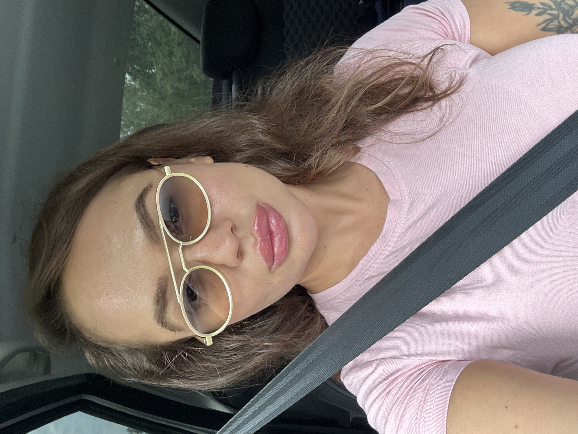 A woman wearing a pink t-shirt and sunglasses is in the driver's seat of a car, with a seatbelt across her chest. She has long brown hair and is looking towards the camera. The background shows the interior of the car and trees outside the window.