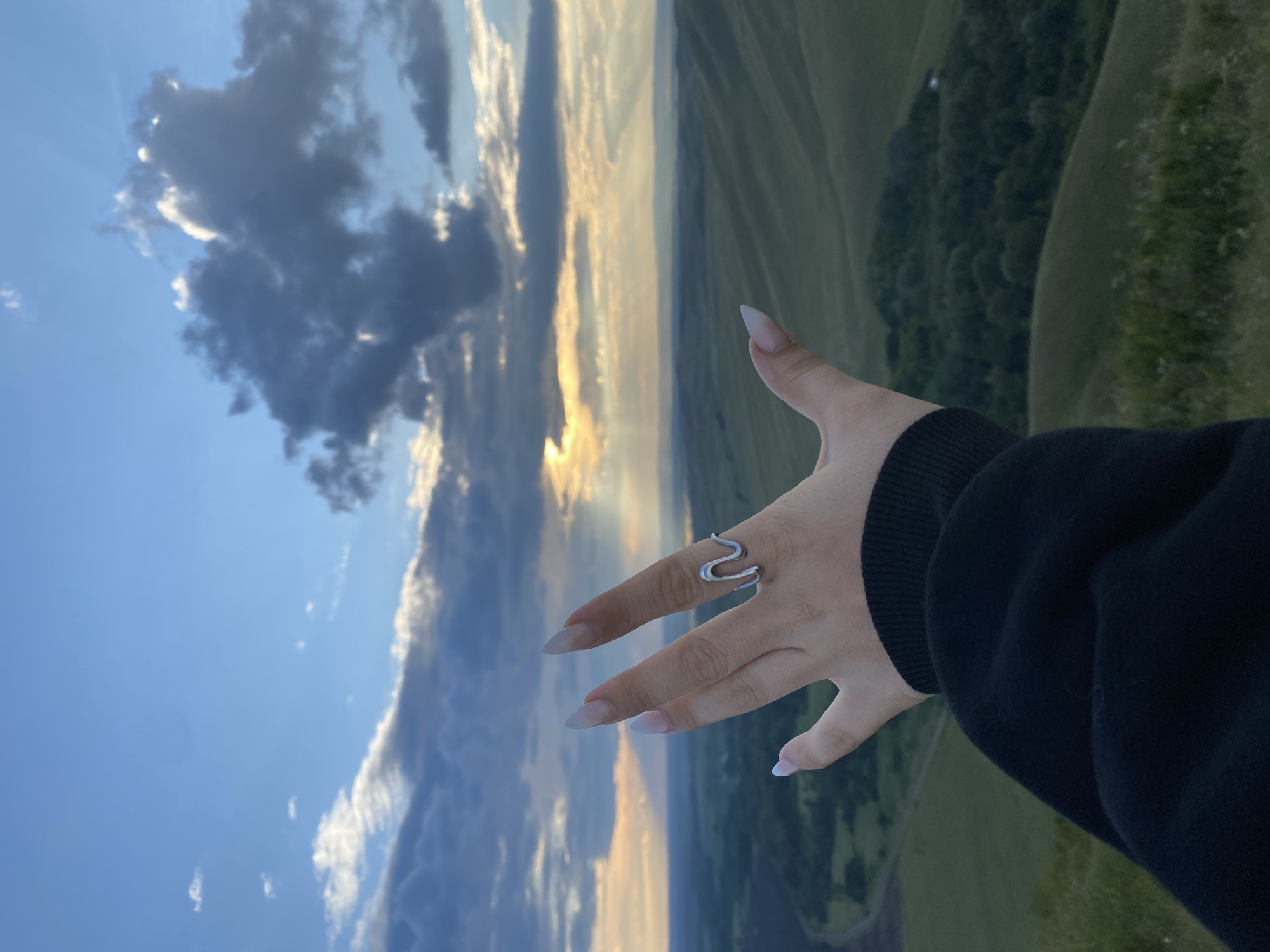 A hand adorned with a silver snake-shaped ring is extended towards a vast, undulating landscape under a dramatic sky. The hand, with long, almond-shaped nails, is positioned in the foreground, partially obscuring the view of the distant terrain. The landscape is characterized by rolling green hills, some sparsely covered with trees and others appearing as open fields. A winding road is faintly visible in the mid-ground. The sky is a blend of deep blue and scattered clouds, with the sun breaking through in a powerful display of crepuscular rays, illuminating the horizon. The overall impression is one of serene natural beauty, with a touch of artistic flair from the ring and the hand's pose. The location is identified as Абзаново, Russia.