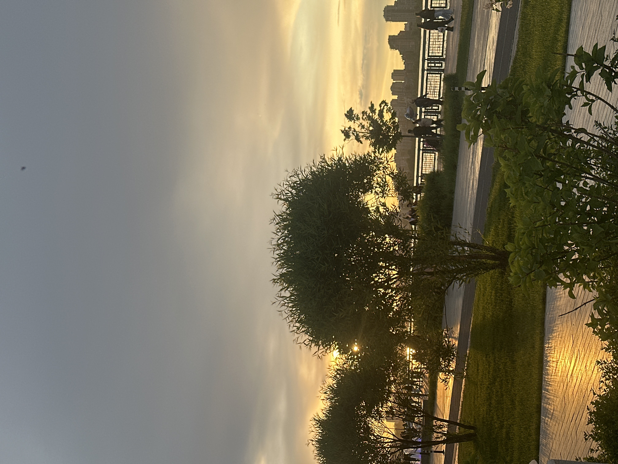 The image shows a park at sunset in Kazan, Russia. Silhouetted figures of people are visible on a walkway lined with a metal railing, suggesting a waterfront area. Tall buildings, likely apartment complexes, are faintly outlined against the bright, golden sky, indicating an urban setting. Green trees and shrubs are in the foreground, with sunlight glinting off a stone walkway, suggesting a well-maintained park area. The sky transitions from a bright golden color near the horizon to a gray hue higher up, indicative of the time of day.