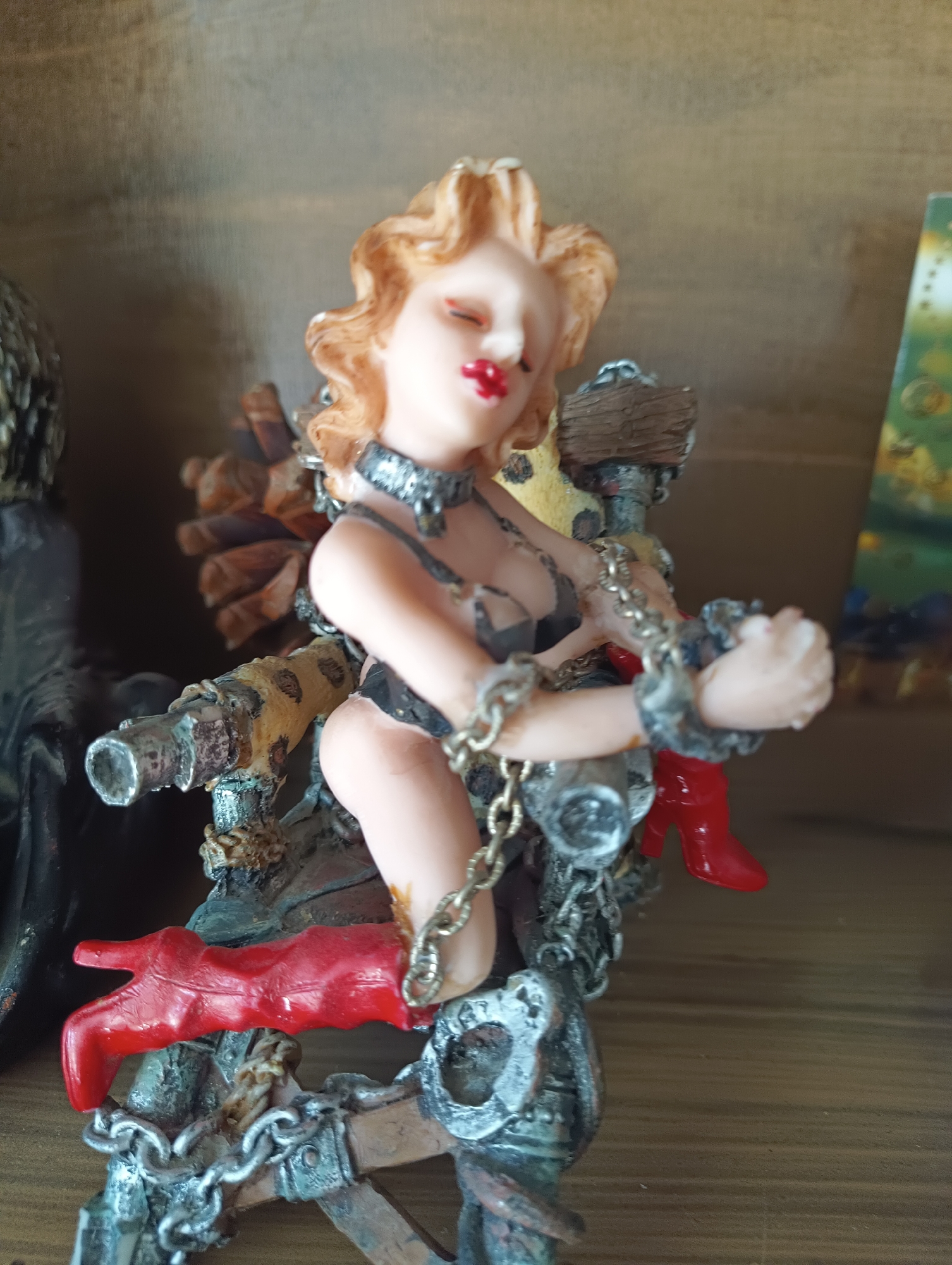 The image shows a figurine of a woman with blonde hair, red lipstick, and a provocative outfit. She is adorned with chains and appears to be in a bondage-themed pose. The figurine is placed on a wooden shelf, with other objects visible in the background. There is no indication or information related to the city of Karnobat, Bulgaria.