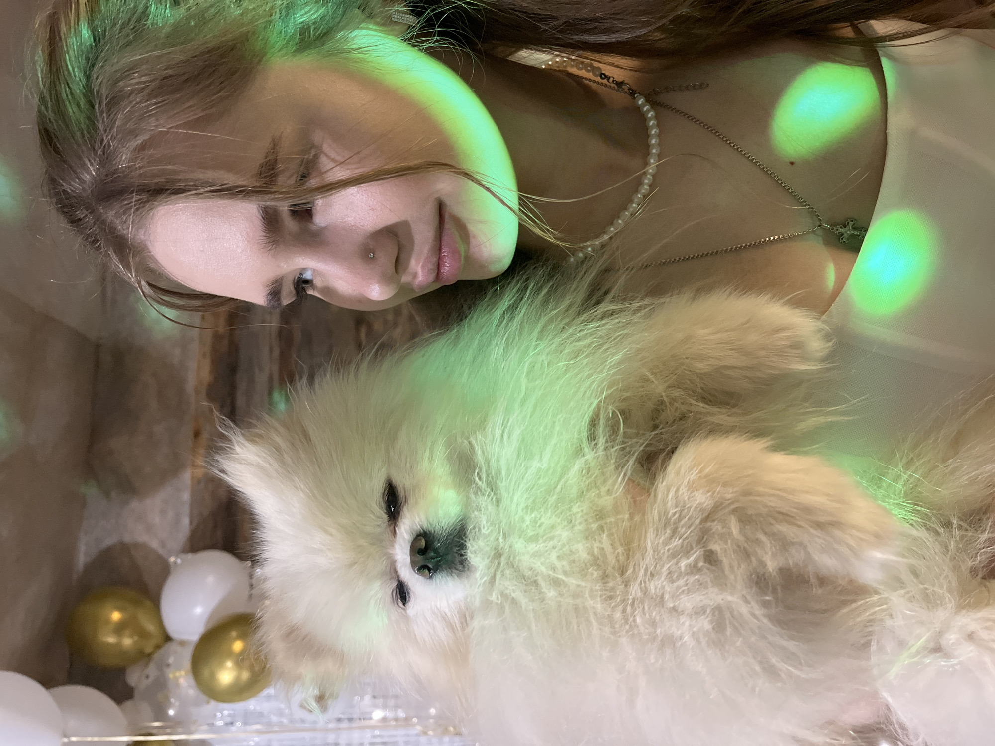 An indoor scene captures a young woman and a fluffy white dog sharing a tender moment. The image is rotated, showing the woman lying on her side, with her head and upper torso visible. She has long brown hair and a gentle, soft smile as she looks towards the dog, which rests close to her. She is wearing a pearl necklace and a silver chain with a cross pendant, and a small stud is visible in her nose.
The dog, likely a Pomeranian, is prominently featured, with its thick, light-colored fur. Its face is visible, with dark eyes and nose. The setting is dimly lit, with distinctive green light projections creating bright, circular patterns on the subjects and the surrounding area, suggesting a festive or party ambiance. In the background, out-of-focus white and gold balloons are discernible, reinforcing the celebratory theme. The moment appears intimate and affectionate, likely taking place in the evening or night. No text is visible in the image. The location context is Izhevsk, Russia.