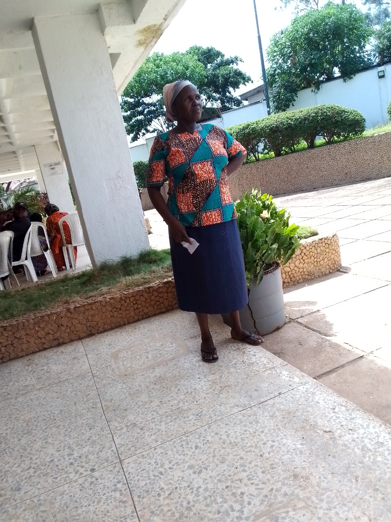 A woman wearing a headscarf, a colorful patterned shirt, and a dark blue skirt stands with her hands on her hips, looking to her left. She is holding a piece of paper in her right hand. She is standing on a paved area in front of a white building with an overhang. In the background, there are trees, bushes, and a white wall. To her left, several people are seated on white chairs under the overhang. The image is taken during the daytime, with sunlight casting shadows on the ground. This scene is consistent with an outdoor waiting area or gathering space in the city of Jos, Nigeria, possibly at a public facility or institution.