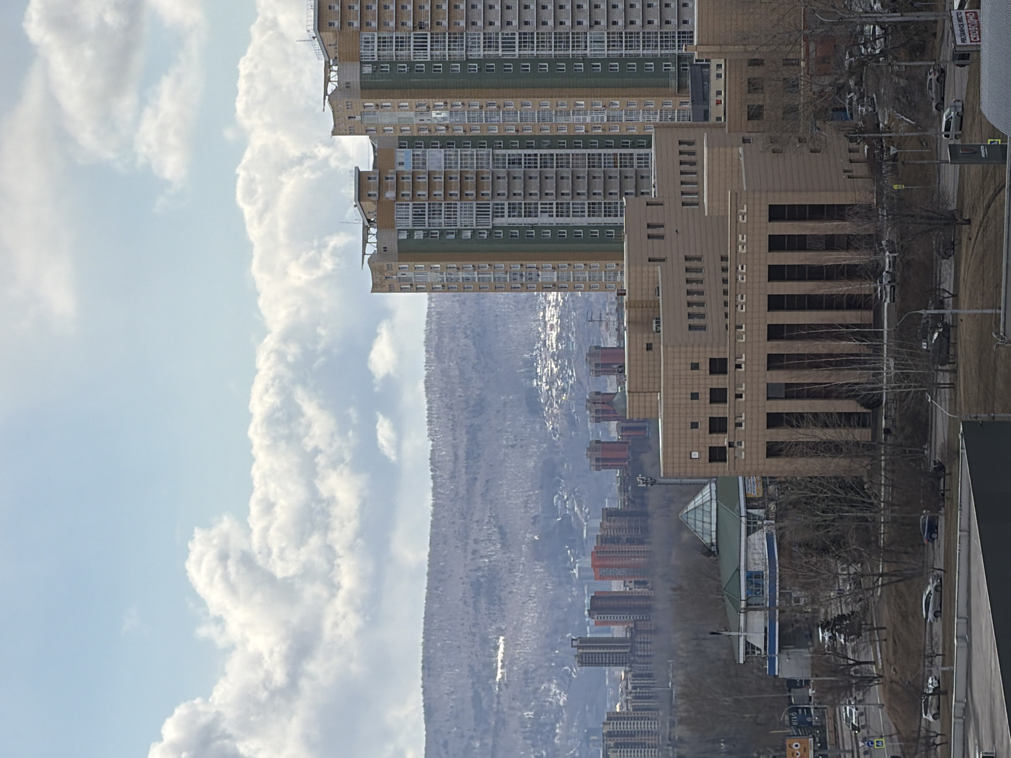The image depicts a cityscape with tall residential and office buildings on the right, stretching upwards into a cloudy sky on the left. In the background, there is a wooded mountain range covered in snow. The photo appears to be taken from a high vantage point, looking down at the city. Along the street, there are sidewalks, roads, trees, cars, and streetlights. The buildings are primarily constructed in light, neutral tones. The image appears to be taken in Krasnoyarsk, Russia.
