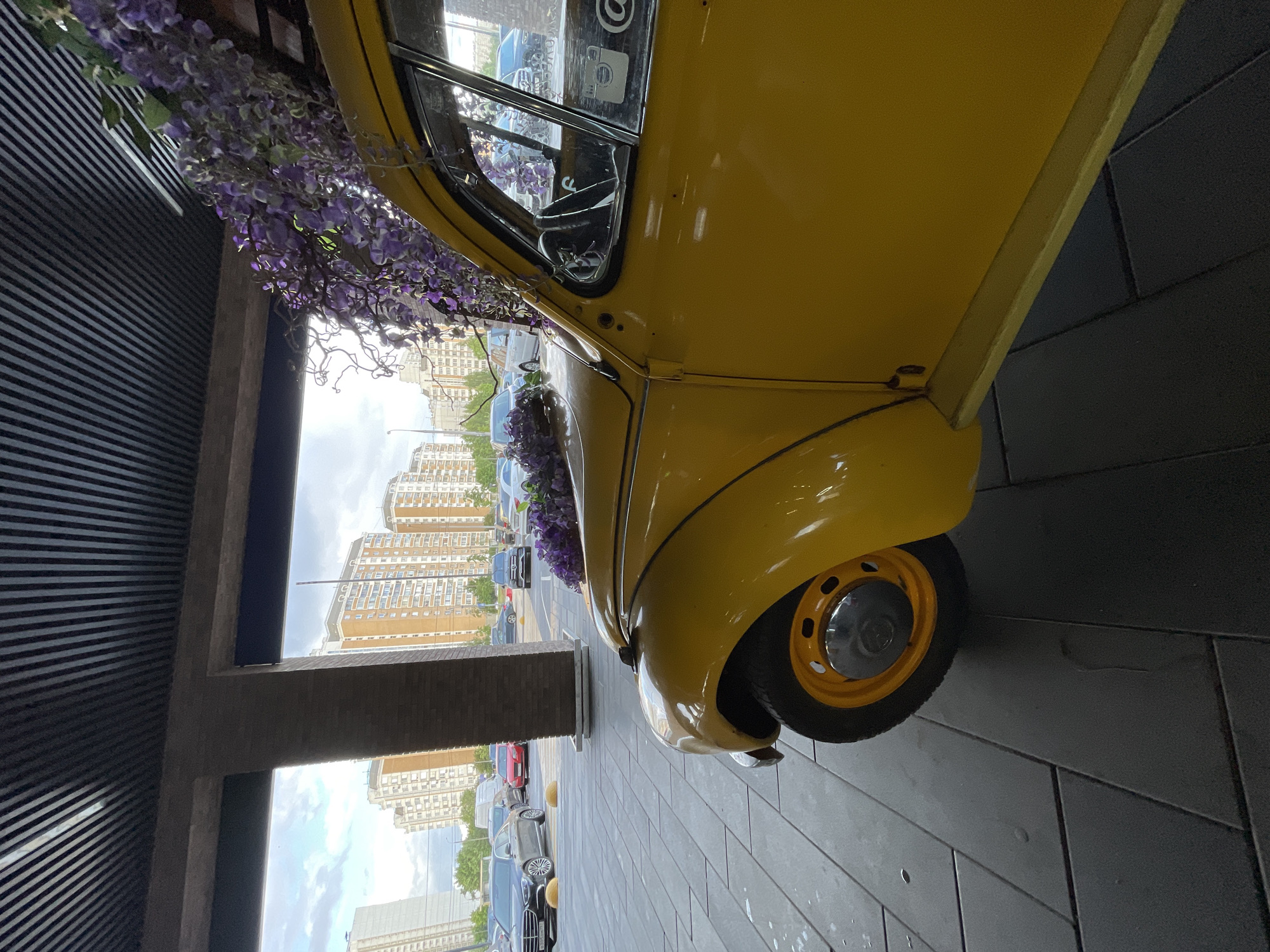 A yellow Volkswagen Beetle is parked on a paved surface under a dark, slatted overhang. The car is facing away from the viewer, with its front-left wheel visible. Purple artificial flowers are draped over the car's hood and extend up and over the roofline. In the background, several multi-story apartment buildings are visible, suggesting an urban environment. A street with parked cars is also present. The scene appears to be in Moscow, Russia, as indicated by the architecture of the buildings and the license plates on the cars. The overall lighting suggests it is daytime, possibly late afternoon. There are no people visible in the image.
