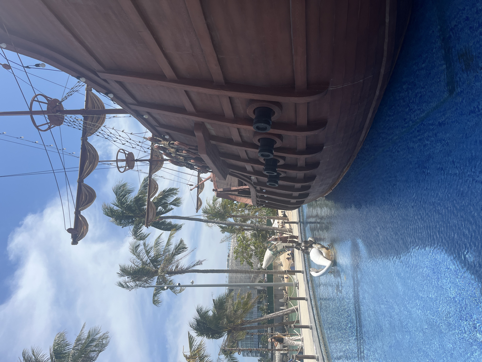 A pirate ship structure is situated next to a blue tiled area. The ship's masts, sails, and cannons are visible. Several palm trees line the area. Water features, including sculptures of swans, are present. A building is visible in the background. People are walking along a walkway nearby. The location appears to be in an area with tropical vegetation.