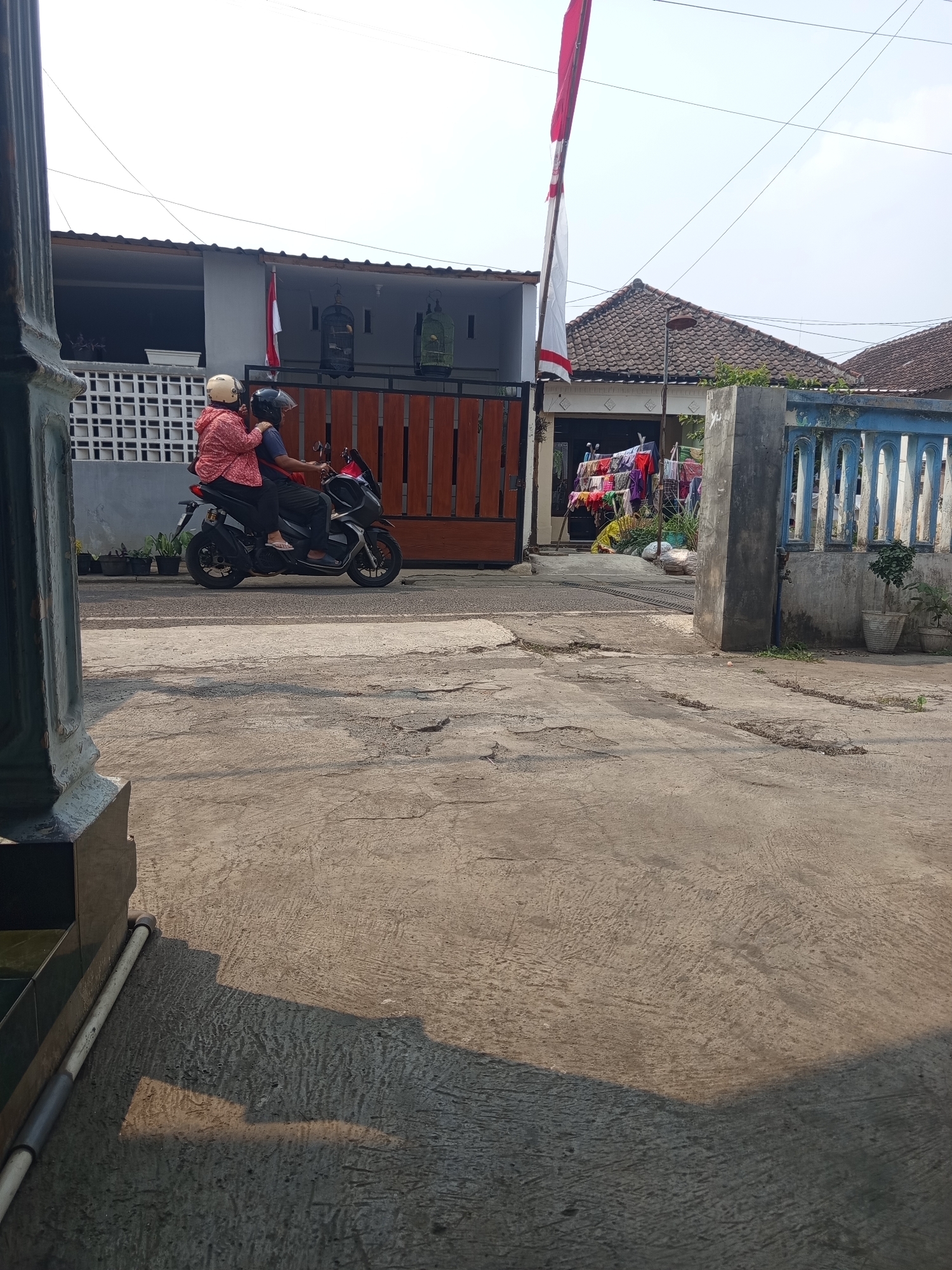 An adult male and an adult female are riding a black motorcycle on a paved road in what appears to be a residential area in the city of Malang, Indonesia. The male is driving, and the female is seated behind him. Both are wearing helmets. The female is wearing a red and white patterned long-sleeved shirt, and the male is wearing a dark blue shirt. They are moving from the foreground to the background, away from the viewer, and are positioned in the center-left of the frame.
The environment consists of a road with a rough, cracked concrete surface. In the background, there are houses with tiled roofs. One house has a prominent wooden gate and a white lattice fence. A red and white Indonesian flag is flying from a pole near the gate. Clothing is seen hanging on a clothesline in front of one of the houses. The sky is bright and clear. The lighting suggests it is daytime, with shadows cast on the road, indicating sunlight. To the far left of the frame, a portion of a dark pillar or structure is visible, partially obscuring the scene. To the right, a blue-colored ornamental fence and potted plants are present. The scene conveys a sense of everyday life in a neighborhood.