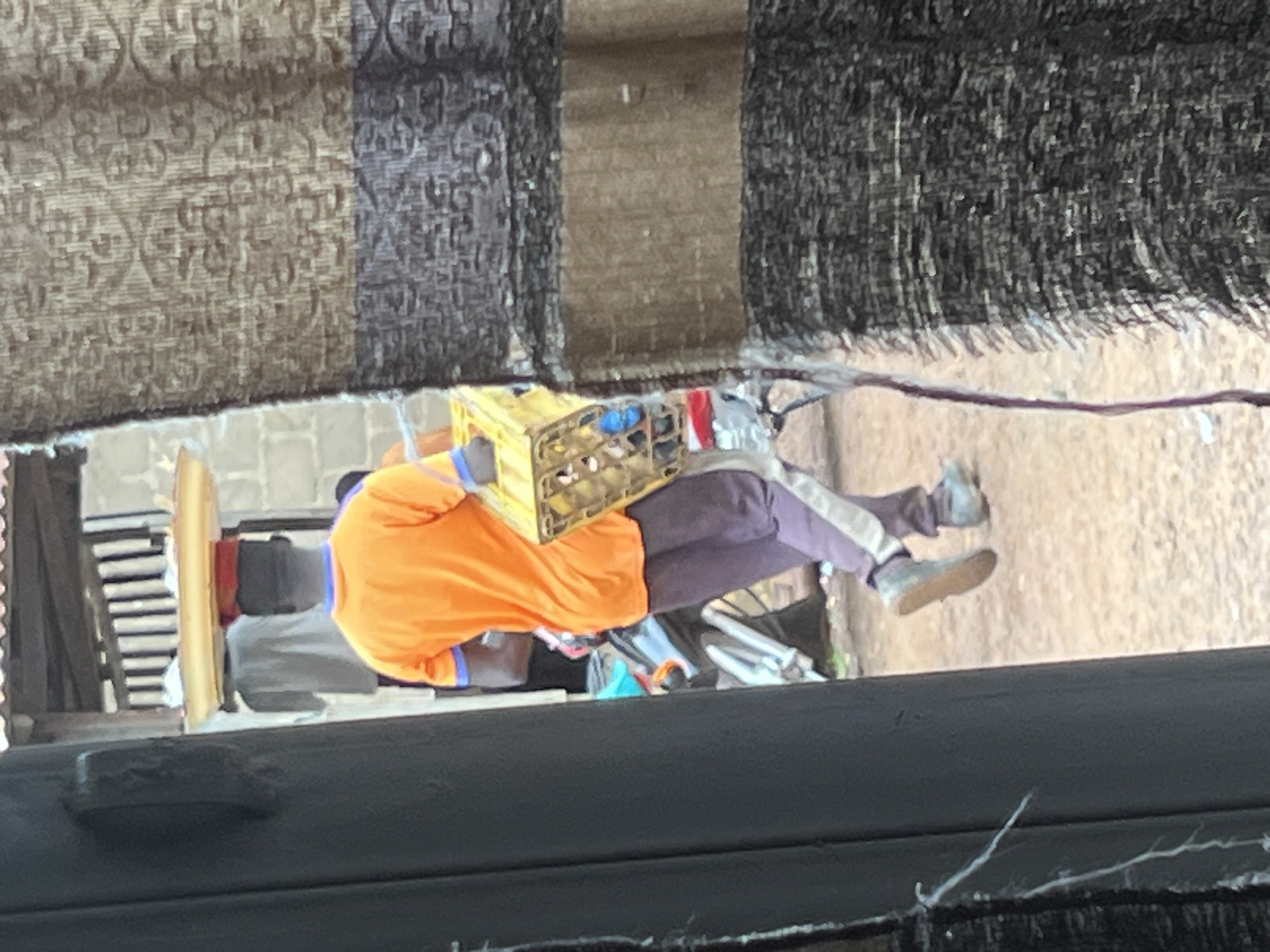 A full-body, rear view shot shows a Black person walking on a dirt path in Durumi, Nigeria. The individual is wearing an orange t-shirt with blue trim at the collar and sleeves, and dark purple athletic pants with a light purple stripe down the side. On their head, they balance a yellow tray with a red circular object. They are carrying a yellow crate of drinks in their right arm. A large, dark gray column is in the left foreground, and a patterned fabric hangs on the right. In the background, a motorcycle is parked behind a brick building with a corrugated metal roof.