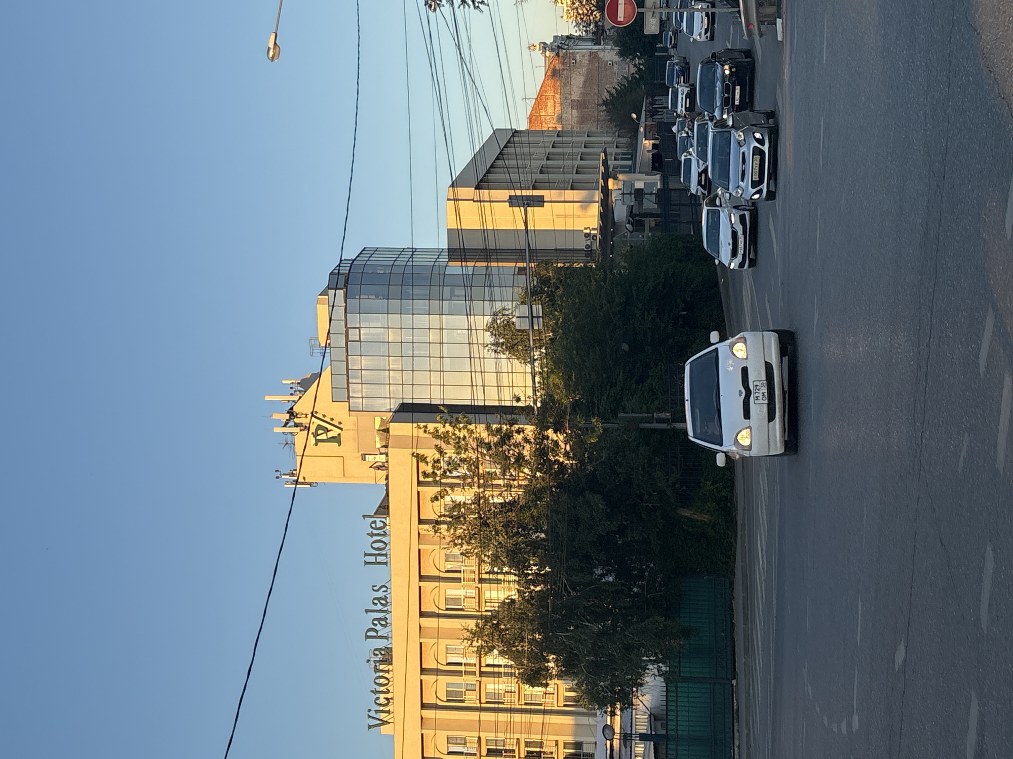 This media file shows a street scene in Astrakhan, Russia, at dusk. The image is oriented vertically. In the foreground, a white car with its headlights on drives down the road. In the middle ground, a modern glass-fronted building stands next to a tan-colored building with the text "Victoria Palas Hotel" prominently displayed on its facade. Trees line the street, and power lines crisscross the sky. In the background, a procession of cars moves along the road, suggesting traffic. The sky is clear and a soft blue, indicating late afternoon or early evening. The overall mood is calm and urban.