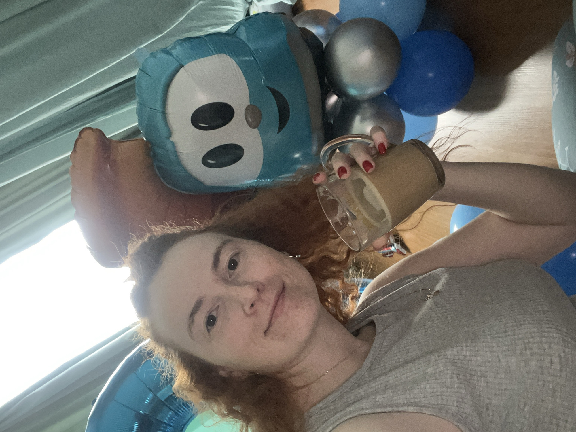 A woman with fair skin and red hair holds a glass mug with a light brown beverage inside, possibly coffee with milk. She is wearing a light gray tank top and has a necklace on. The woman is positioned amongst several balloons, including a large blue cartoon character balloon and several blue and silver balloons. The image appears to be taken indoors, possibly a residence in Chaikovsky, Russia, given the prompt's constraints. A window with light-colored curtains is visible in the background. The photo appears to be a selfie.