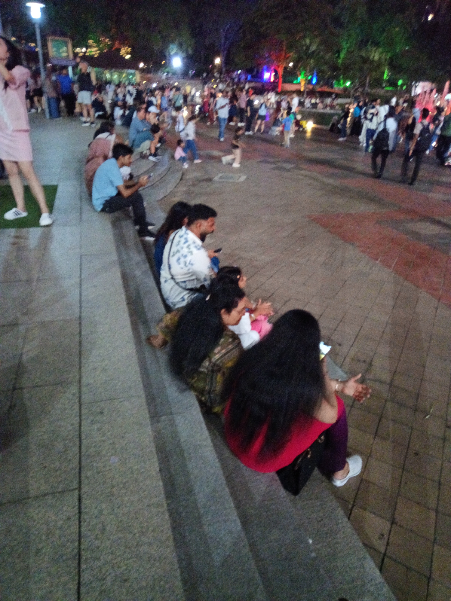 The image captures a nighttime scene at a public gathering, possibly a park or a plaza, in Kuala Lumpur, Malaysia. The ground is paved with stone tiles and concrete steps. In the foreground, several individuals are seated on the steps, facing away from the camera. One person in the front wears a red top and dark purple pants, with their left hand resting on their lap. Behind them, another person is partially visible, wearing a white and blue patterned shirt and dark pants, also seated. Further back, more people are seated on the steps, some appearing to be engaged with their mobile phones.
The background shows a crowd of people milling about in a dimly lit area, with trees and colorful lights illuminating the scene. The lights suggest a festive or event-like atmosphere. The overall impression is one of a casual outdoor event in an urban setting, likely a popular spot for evening gatherings in Kuala Lumpur.